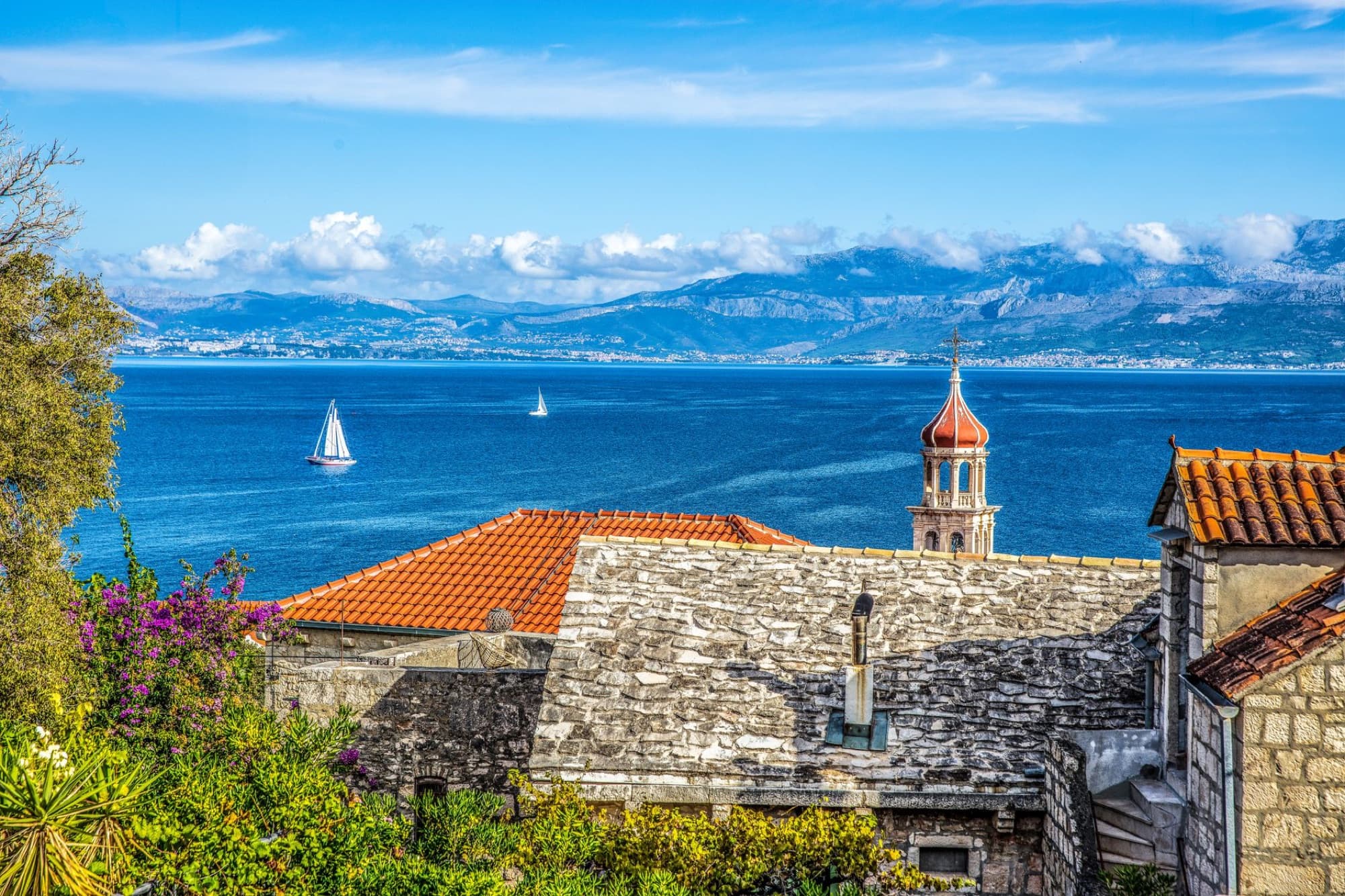 Charming Adriatic coastal village with sailboats and distant mountains under a vivid blue sky.