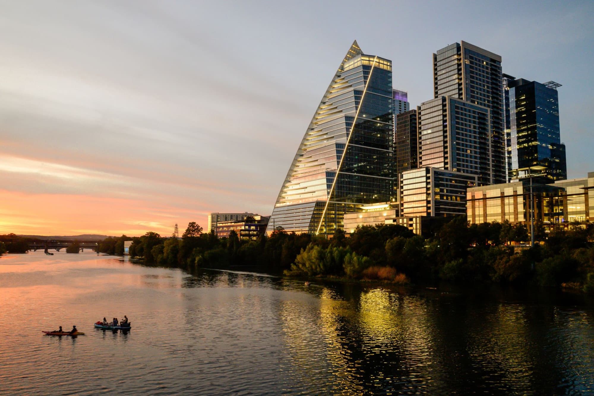 Dramatic sunset over the Austin skyline with reflections on the river and visible kayakers.