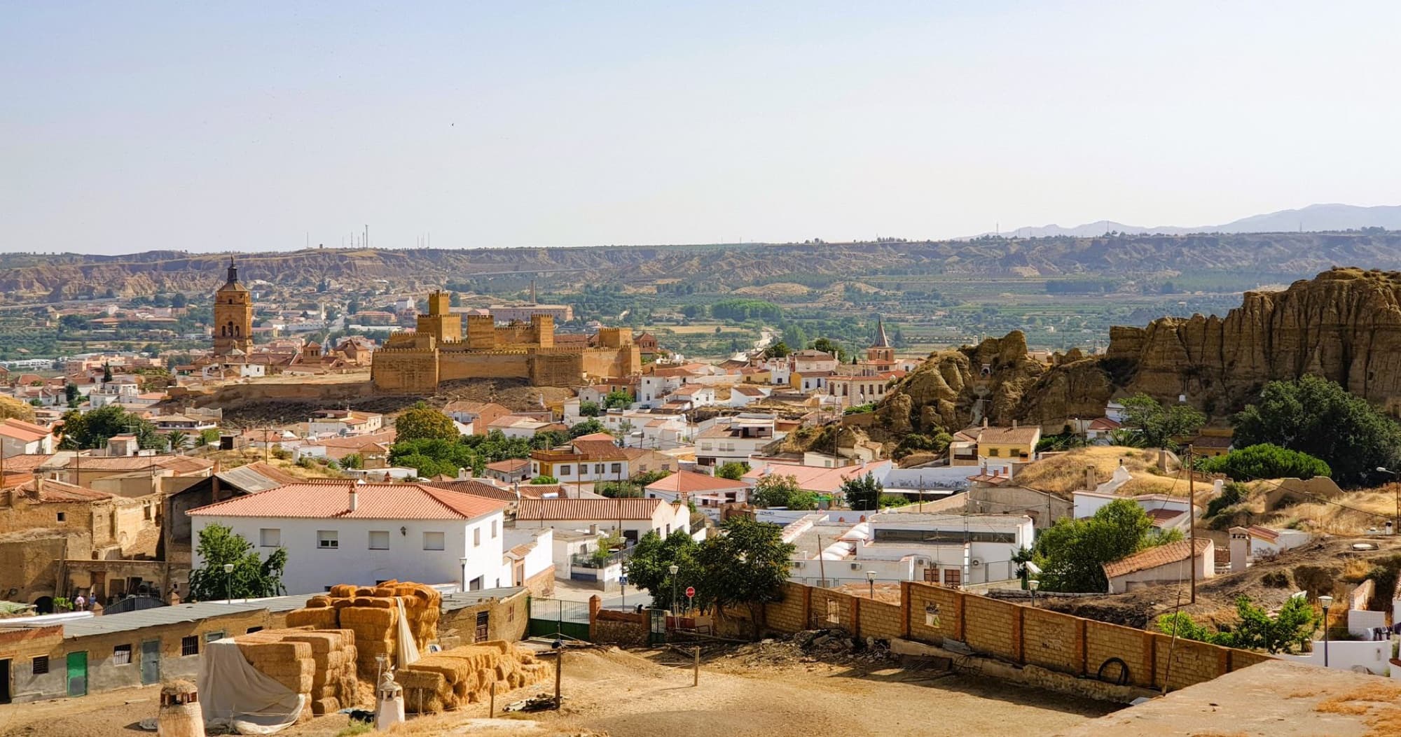 Aerial view of Guadix town with its historic castle and rugged landscape under a clear summer sky.