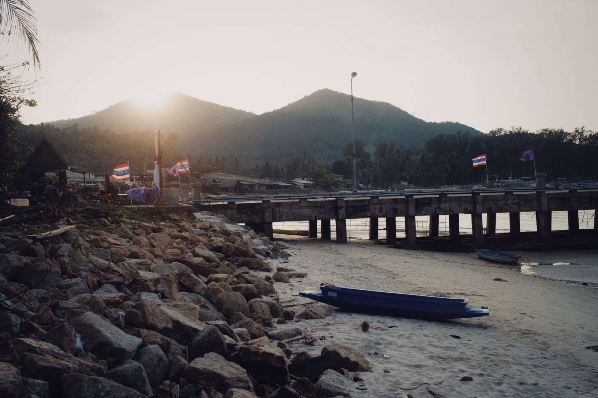Picturesque beach with rocky shore, pier, and mountain view at sunset with Thai flags visible.
