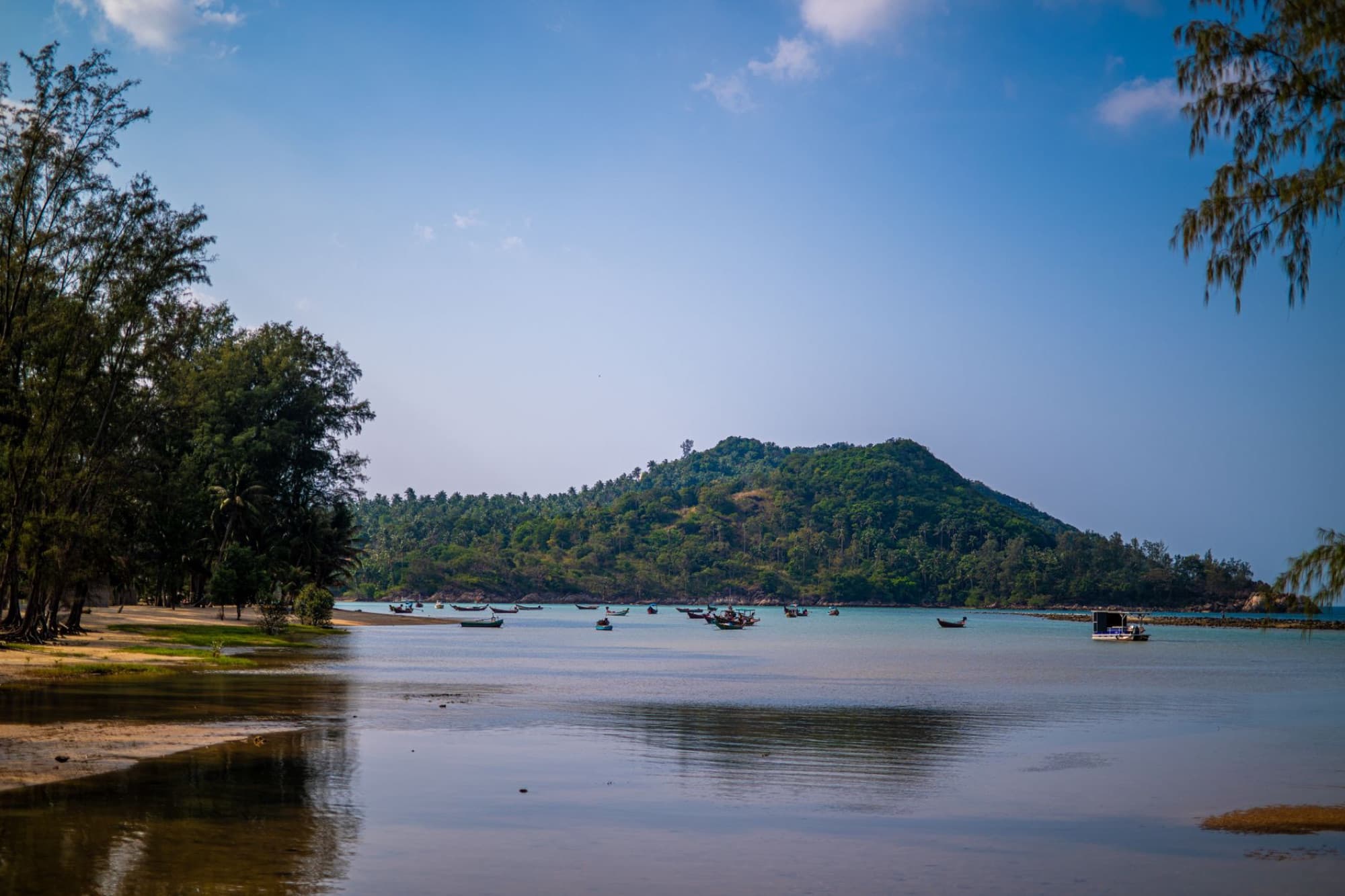 Explore a tranquil tropical beach with scattered boats, lush green island backdrop, and calm waters.