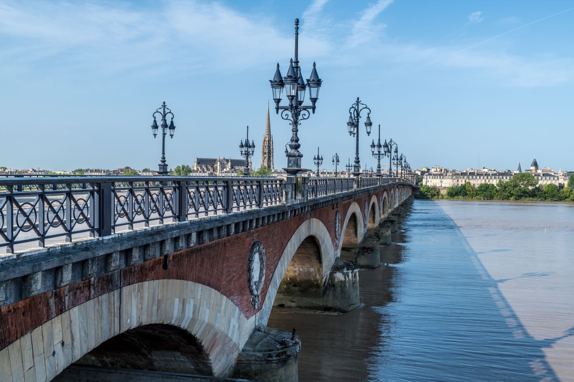 Scenic view of the iconic Pont de Pierre bridge in Bordeaux under a blue sky, reflecting on the calm waters of the Garonne River.