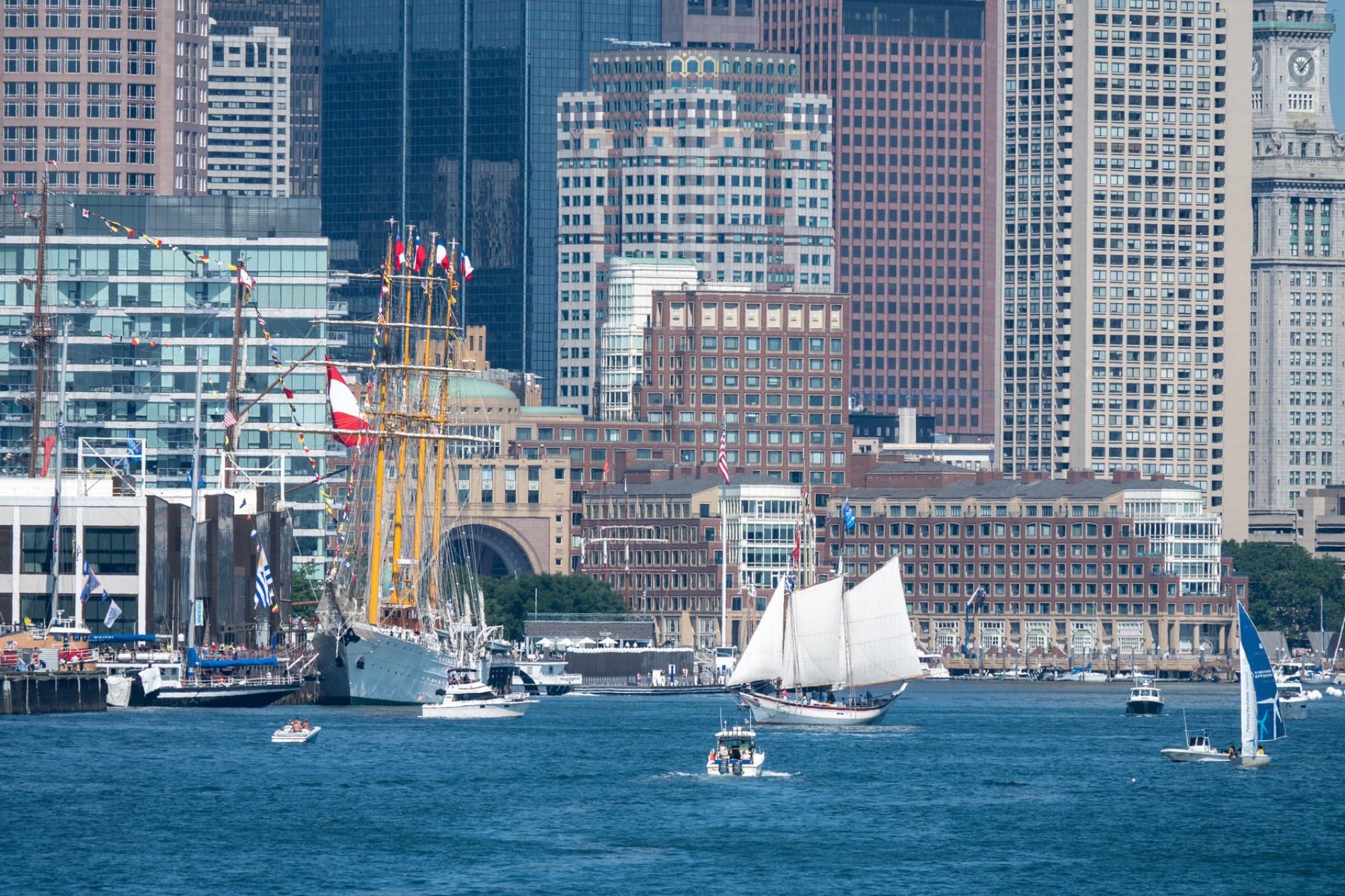 Sailboats navigate Boston Harbor under a summer sky, set against the iconic city skyline.