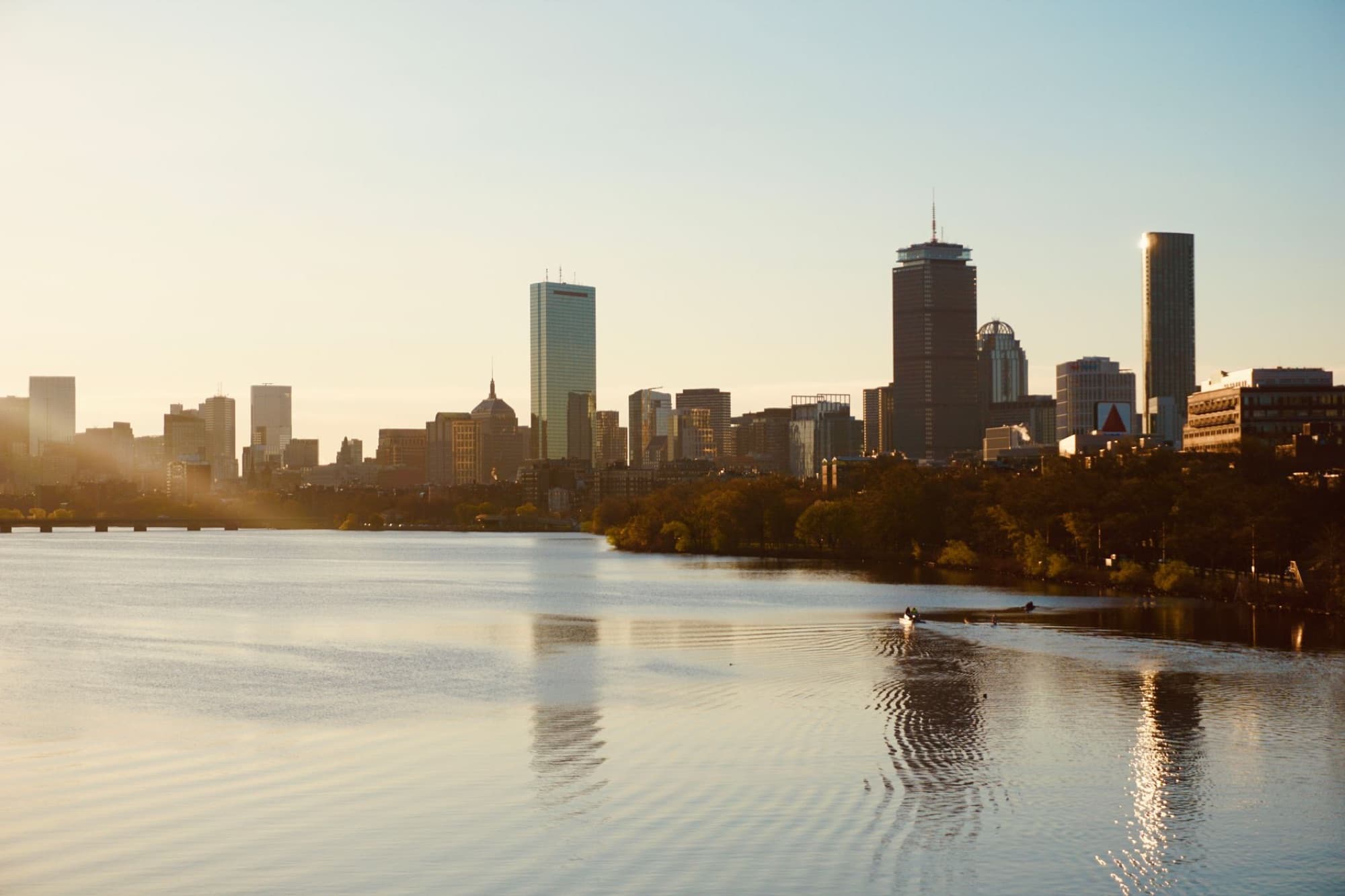 Serene view of Boston skyline at sunset from Charles River, highlighting cityscape and tranquil water.