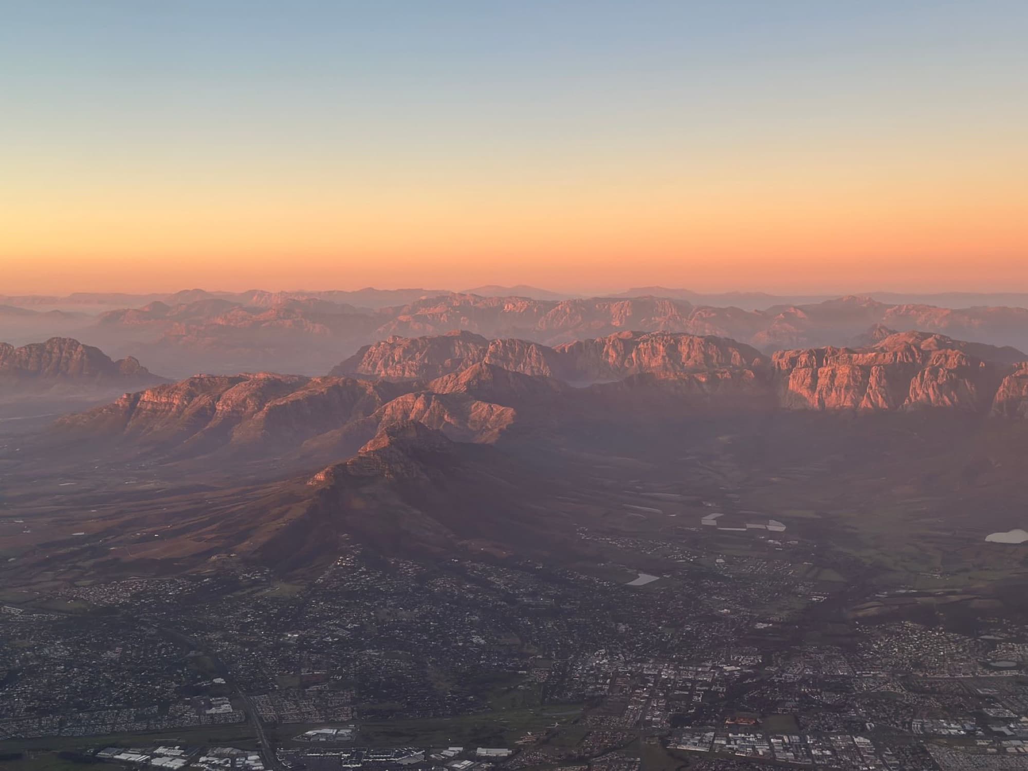 Breathtaking aerial view of rocky mountains with glowing sunset hues and distant cityscape below.