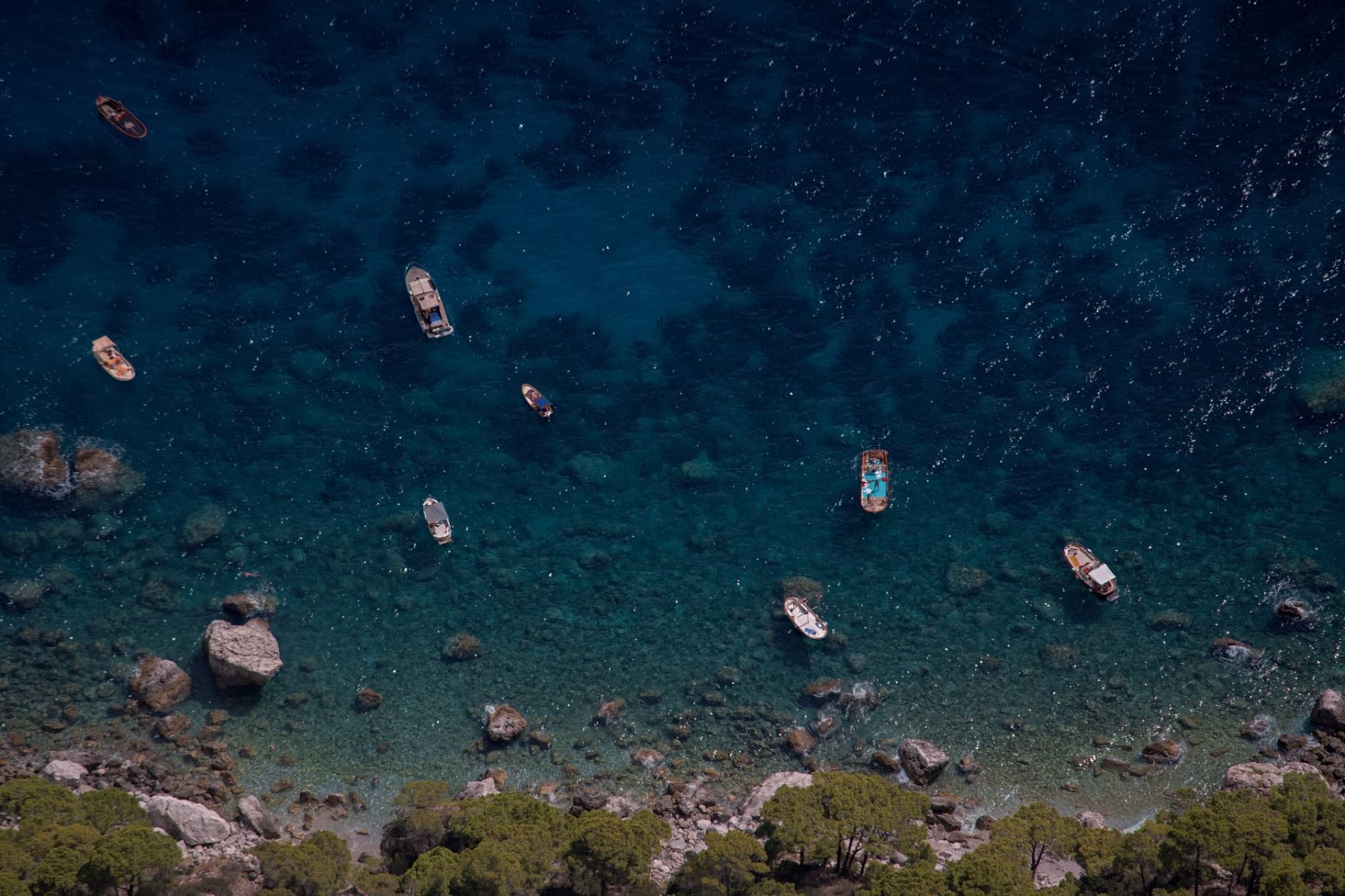 Aerial view of boats floating on the turquoise waters near the rocky coastline of Capri, Italy.