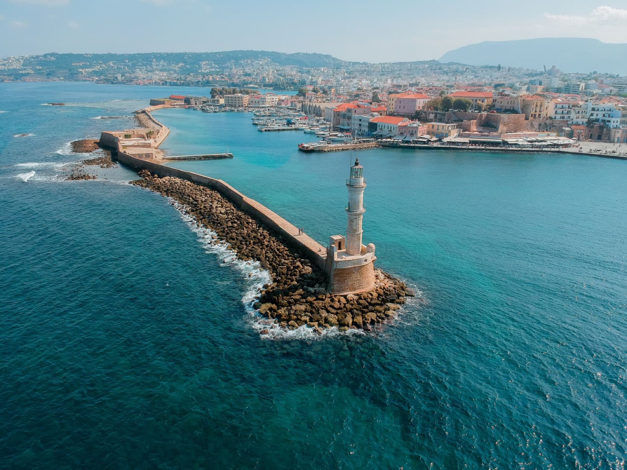 Drone capture of Chania's historic Venetian harbor, showcasing the iconic lighthouse against a scenic coastal backdrop.