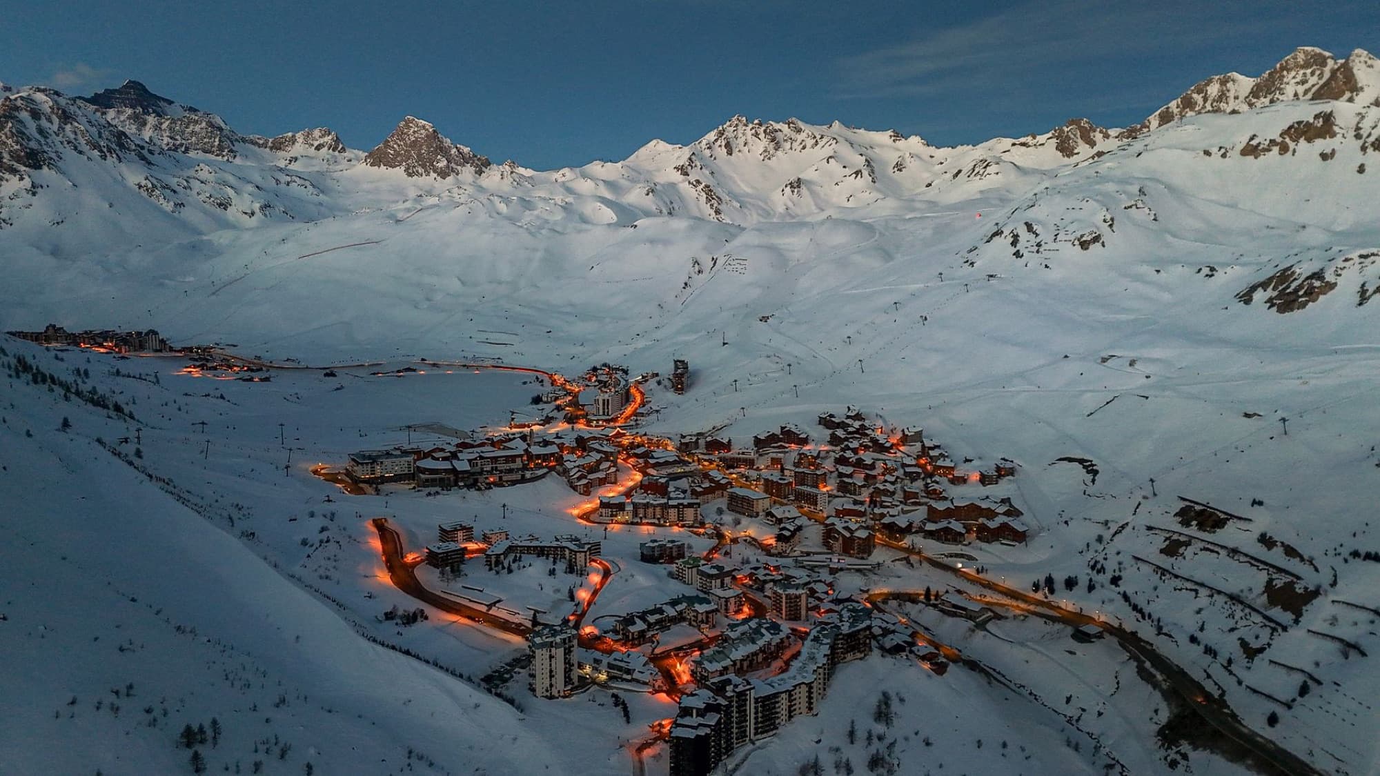 Breathtaking aerial view of a snowy mountain village illuminated at dusk, surrounded by majestic peaks.