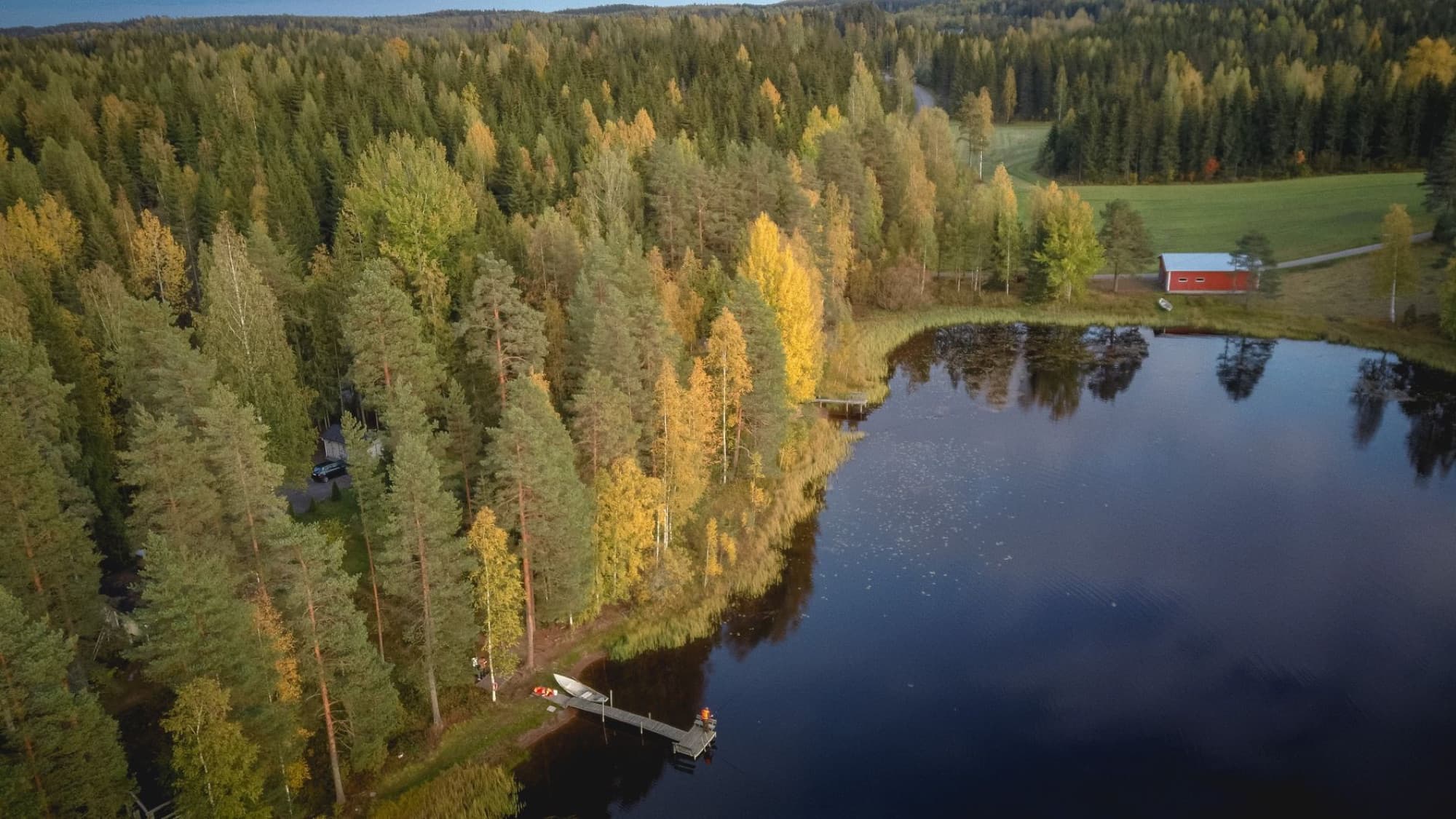Scenic aerial view of a lake and autumn forest in Finland featuring colorful foliage and a red cabin.