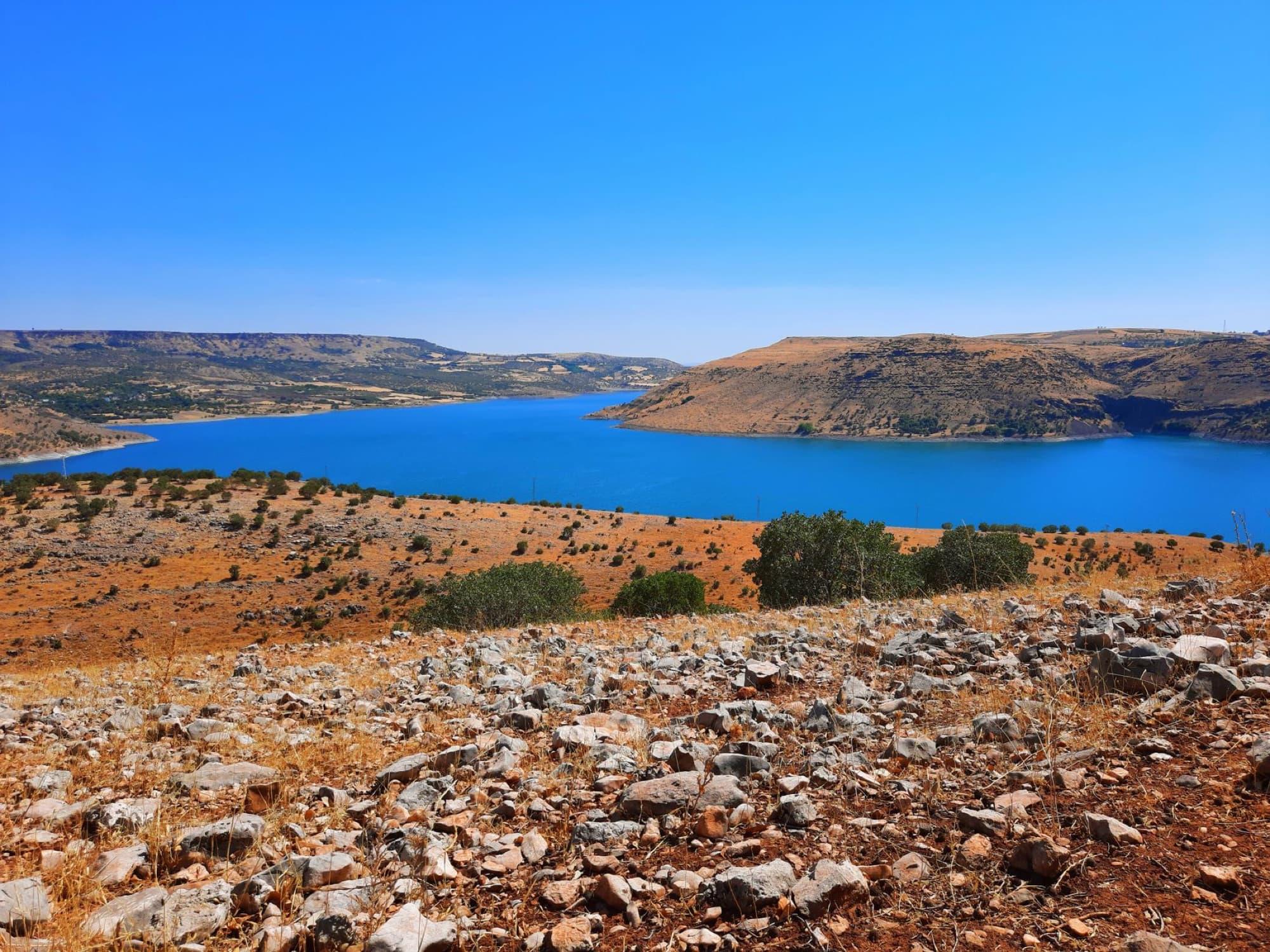 Scenic view of a blue lake surrounded by rocky hills and dry landscape under a clear summer sky.