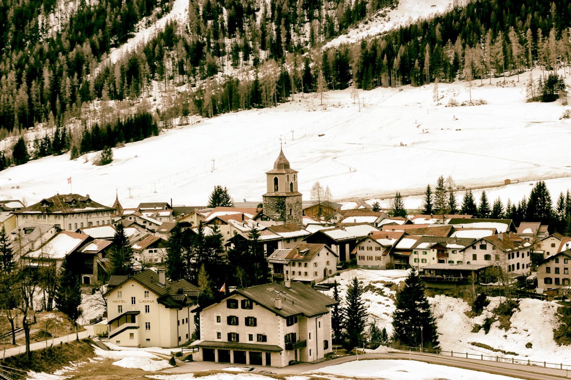 Scenic aerial view of a snowy Alpine village surrounded by mountains and trees in winter.