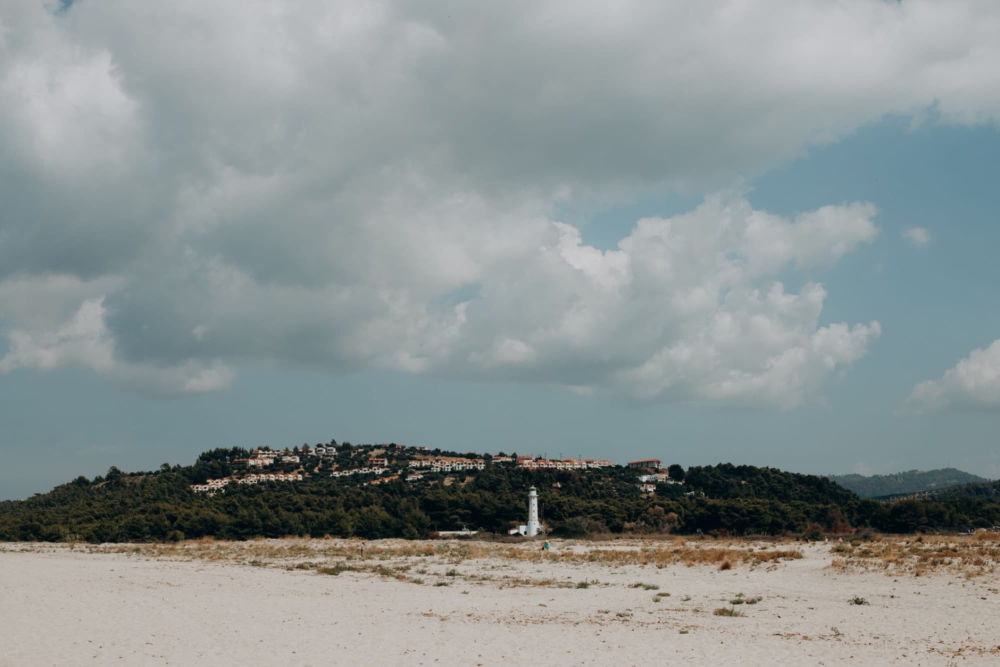 Scenic Greek beach landscape with lighthouse and hillside village under fluffy clouds.