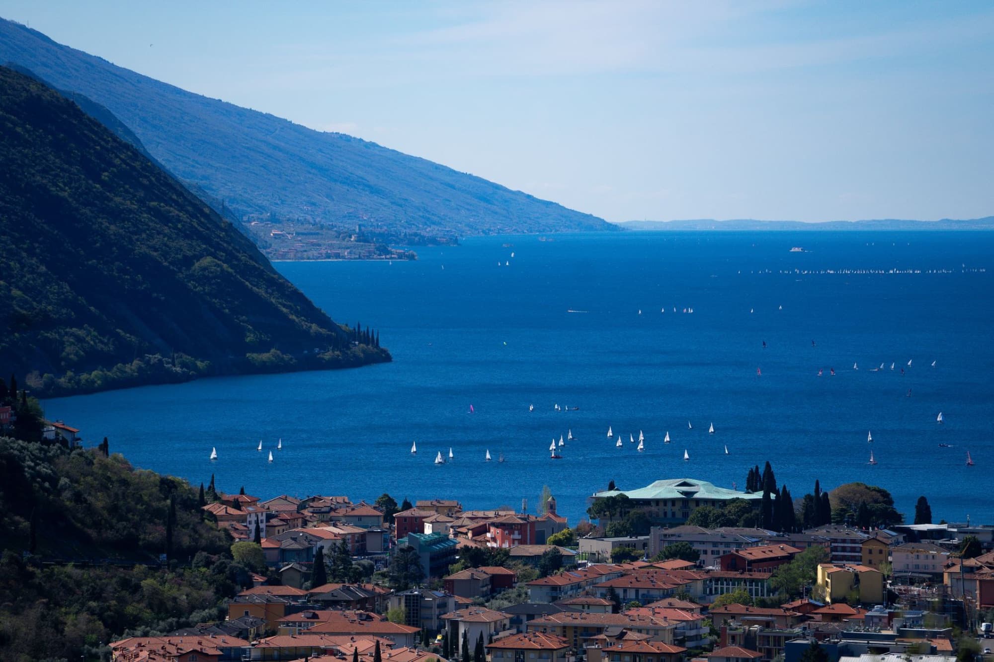 Aerial view of Lake Garda with numerous sailboats on a sunny day, surrounded by mountains.