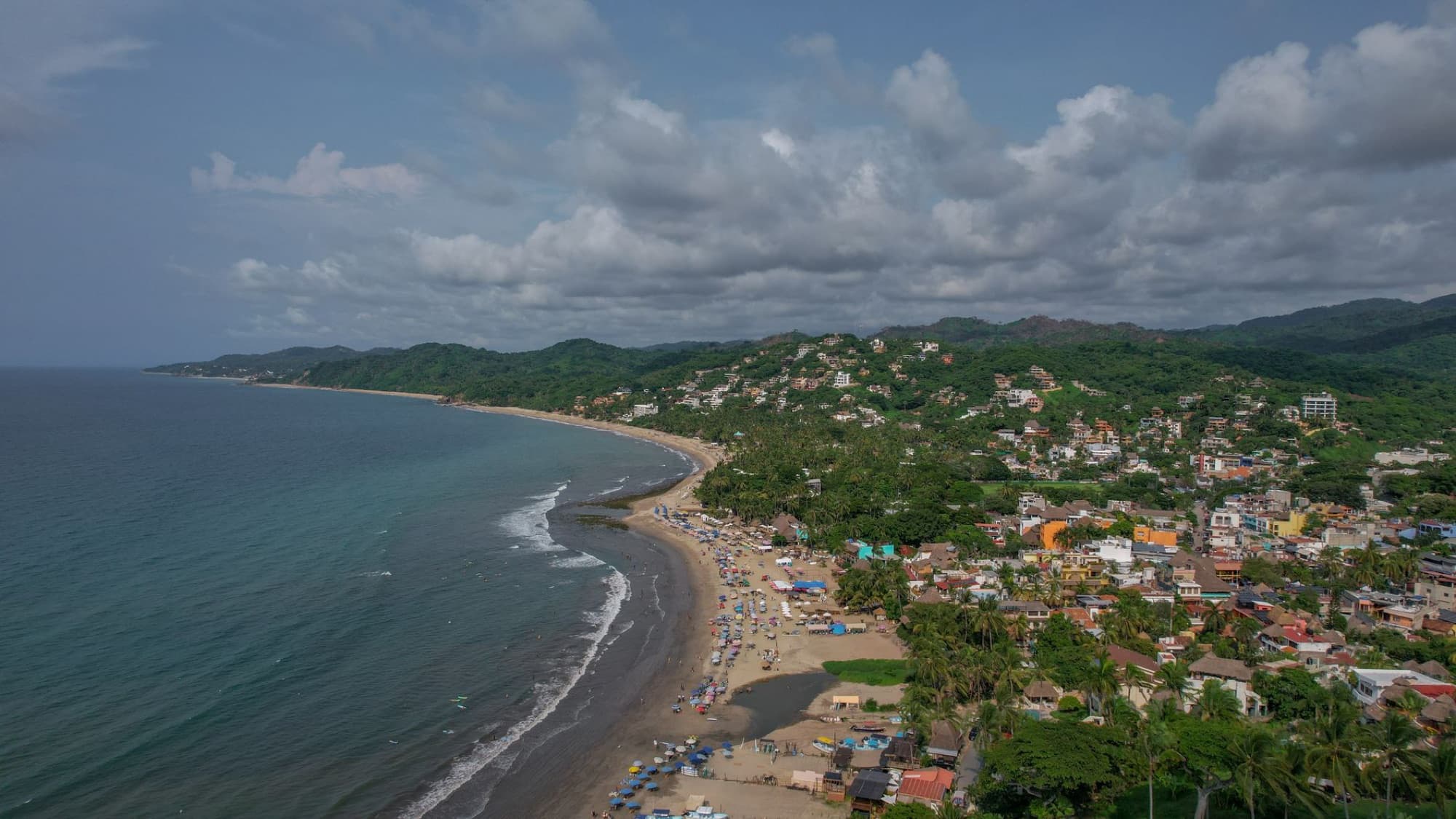A scenic aerial view of a coastal town nestled by lush green hills and a sandy beach along the ocean.