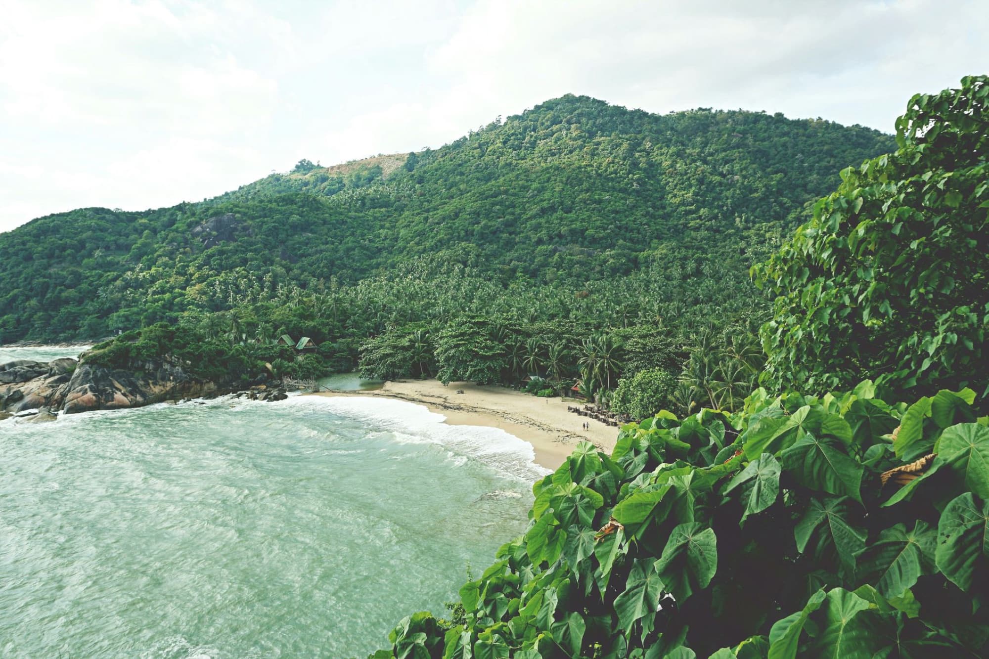 A picturesque tropical beach surrounded by lush green hills in Koh Phangan, Thailand.