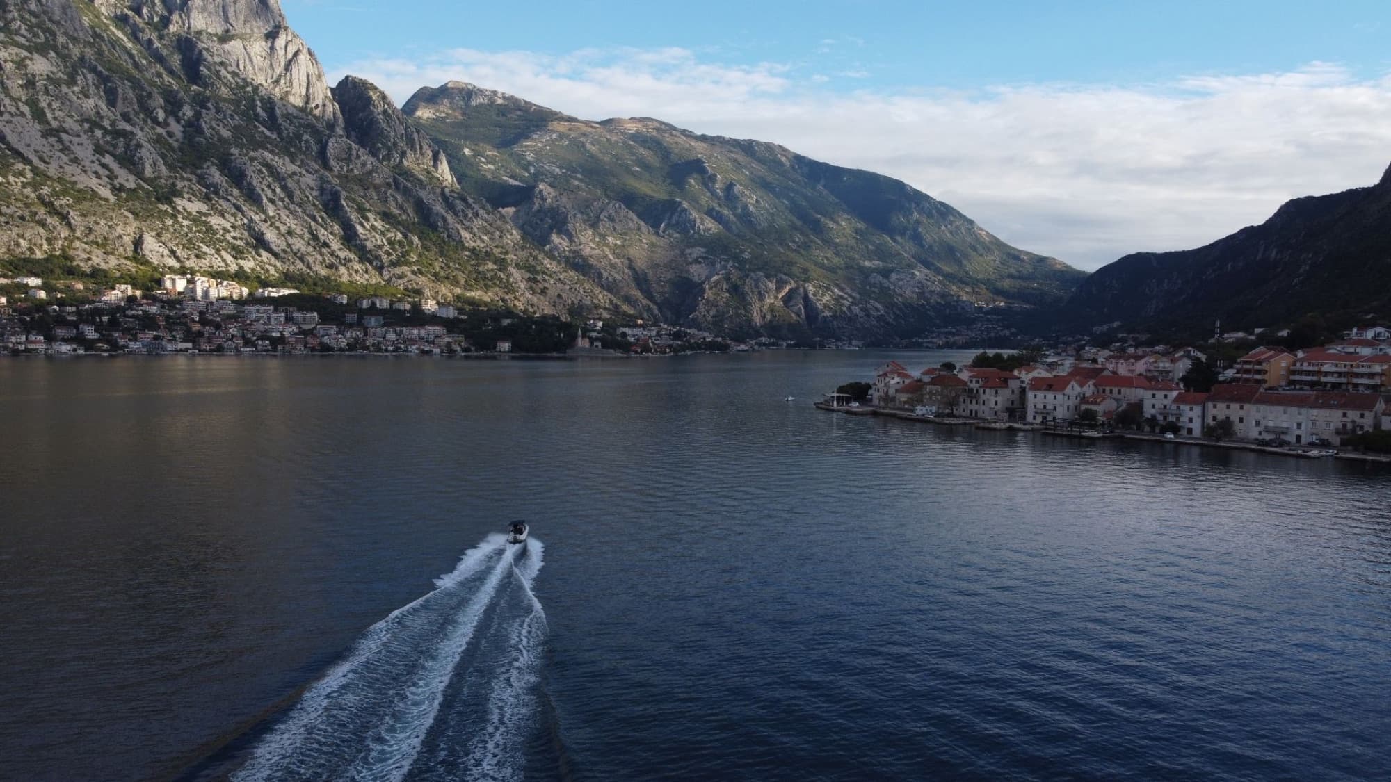 Scenic aerial photograph of Kotor Bay, Montenegro. Features a motorboat gliding through the water with majestic mountains in the background.