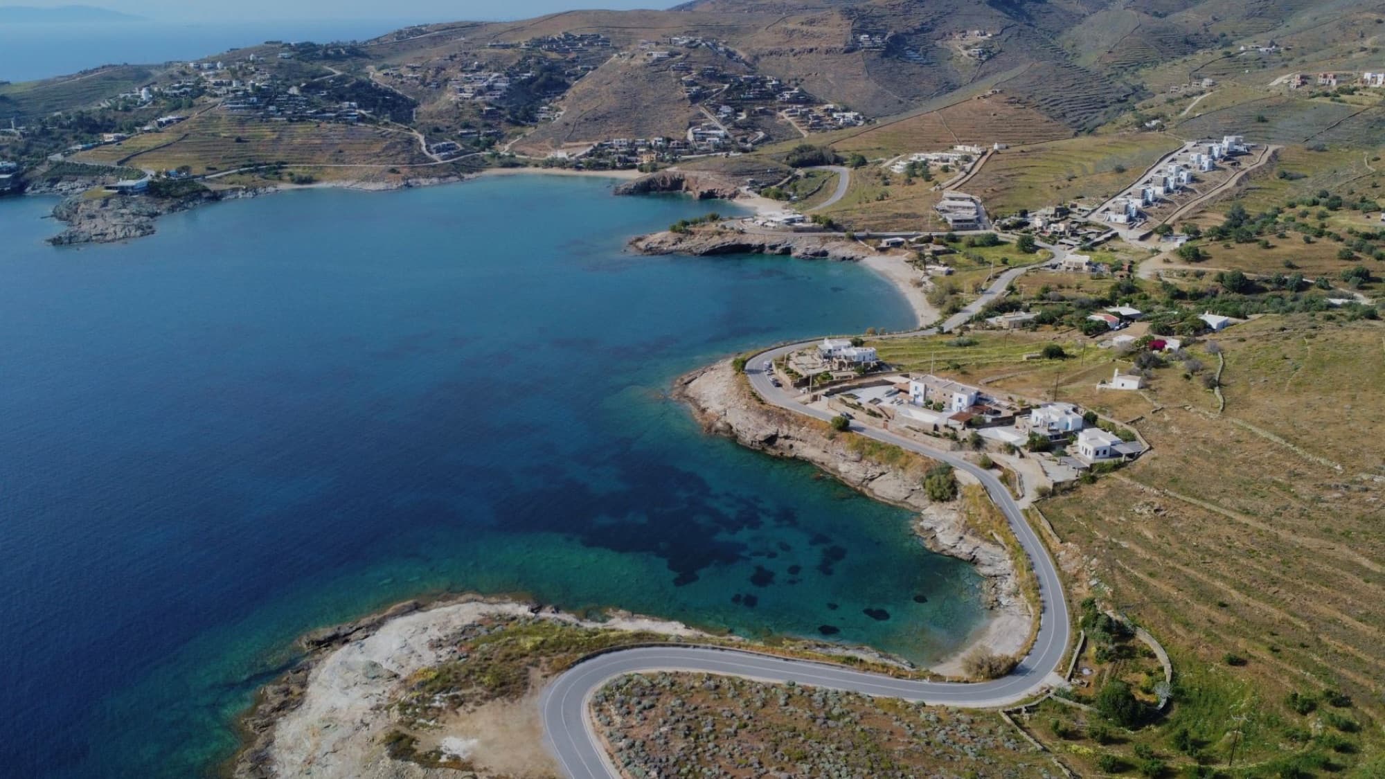 Scenic aerial view of a coastal road and landscape on a Greek island with clear blue waters and rugged hillsides.