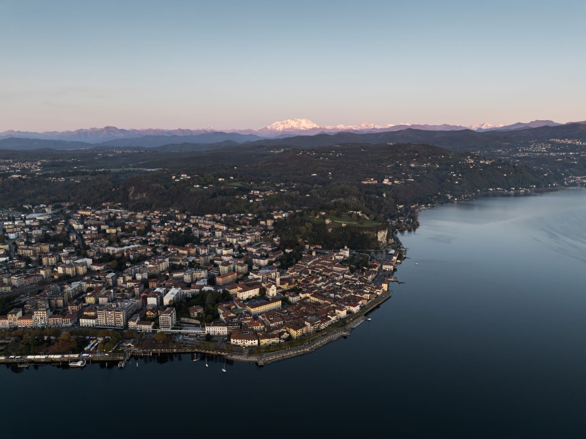 Aerial panorama of Arona, Italy by Lake Maggiore at dusk, with scenic mountains in the background.
