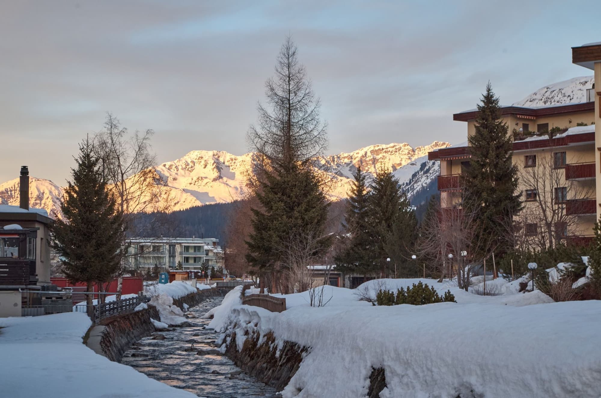 Stunning winter mountain village view with snowy trees and river at sunset.