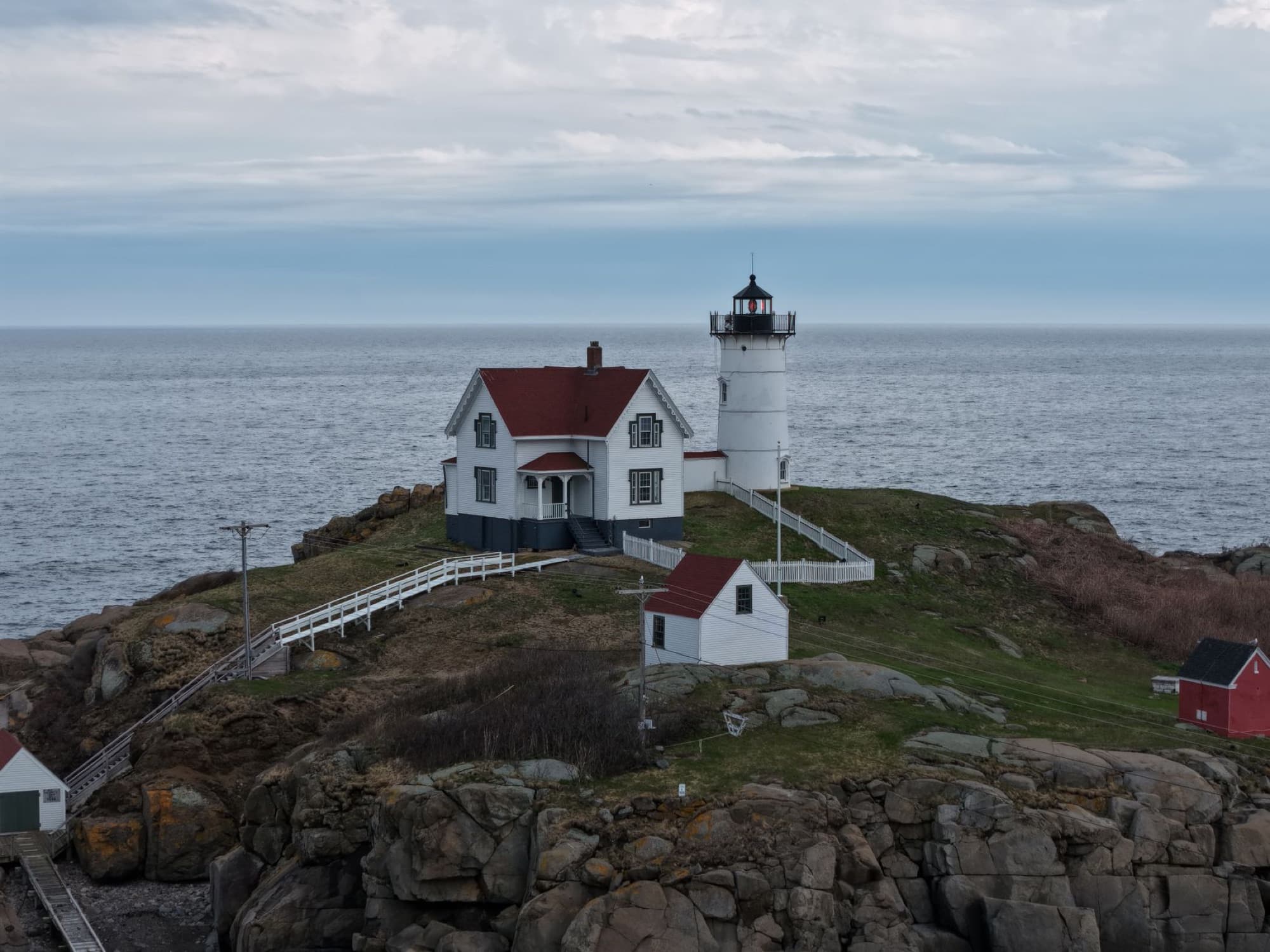 Drone shot capturing the iconic Nubble Lighthouse on a rocky Maine coast under a cloudy sky.