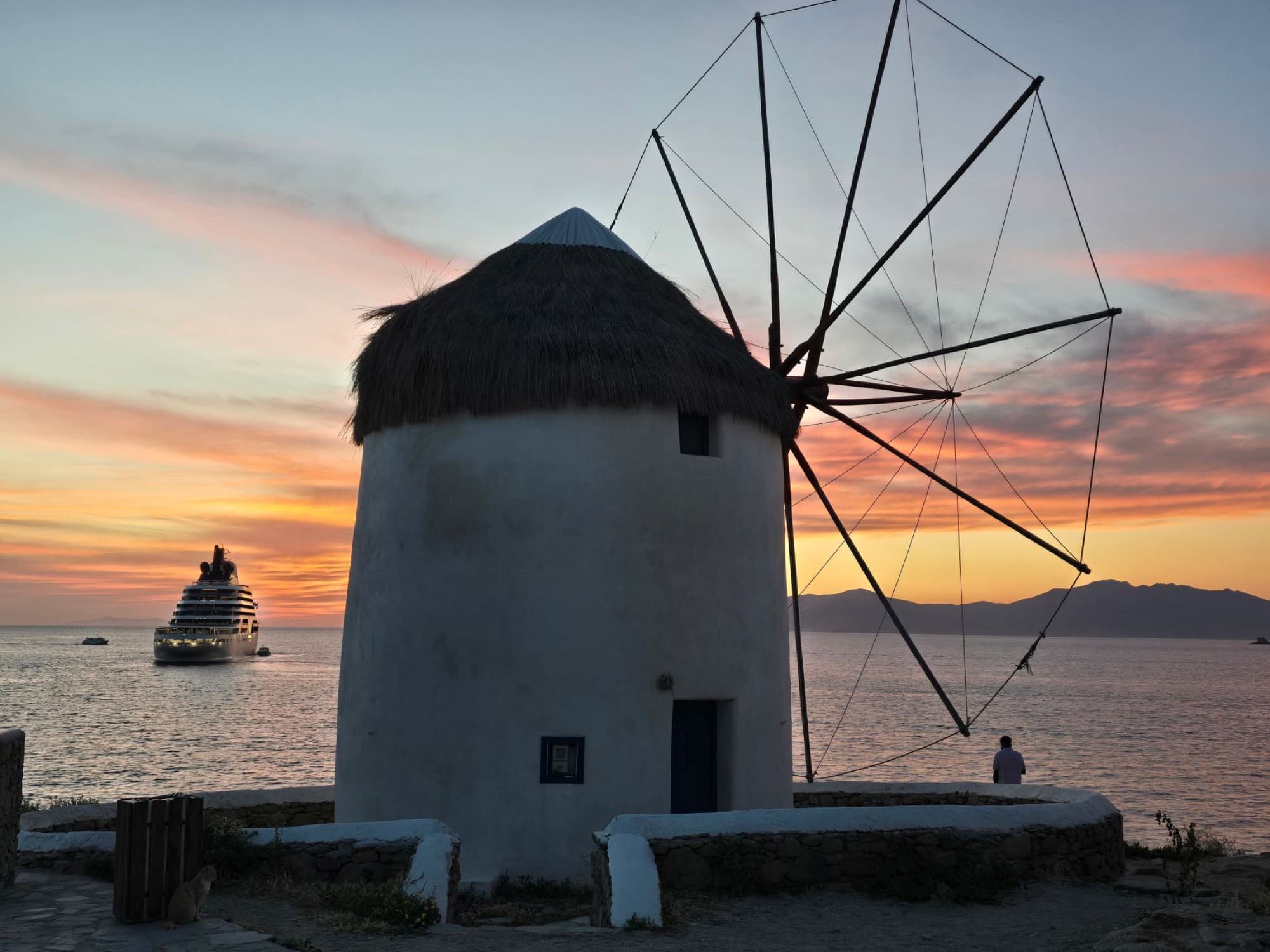 A scenic sunset view of a traditional windmill in Mykonos with a cruise ship in the background.