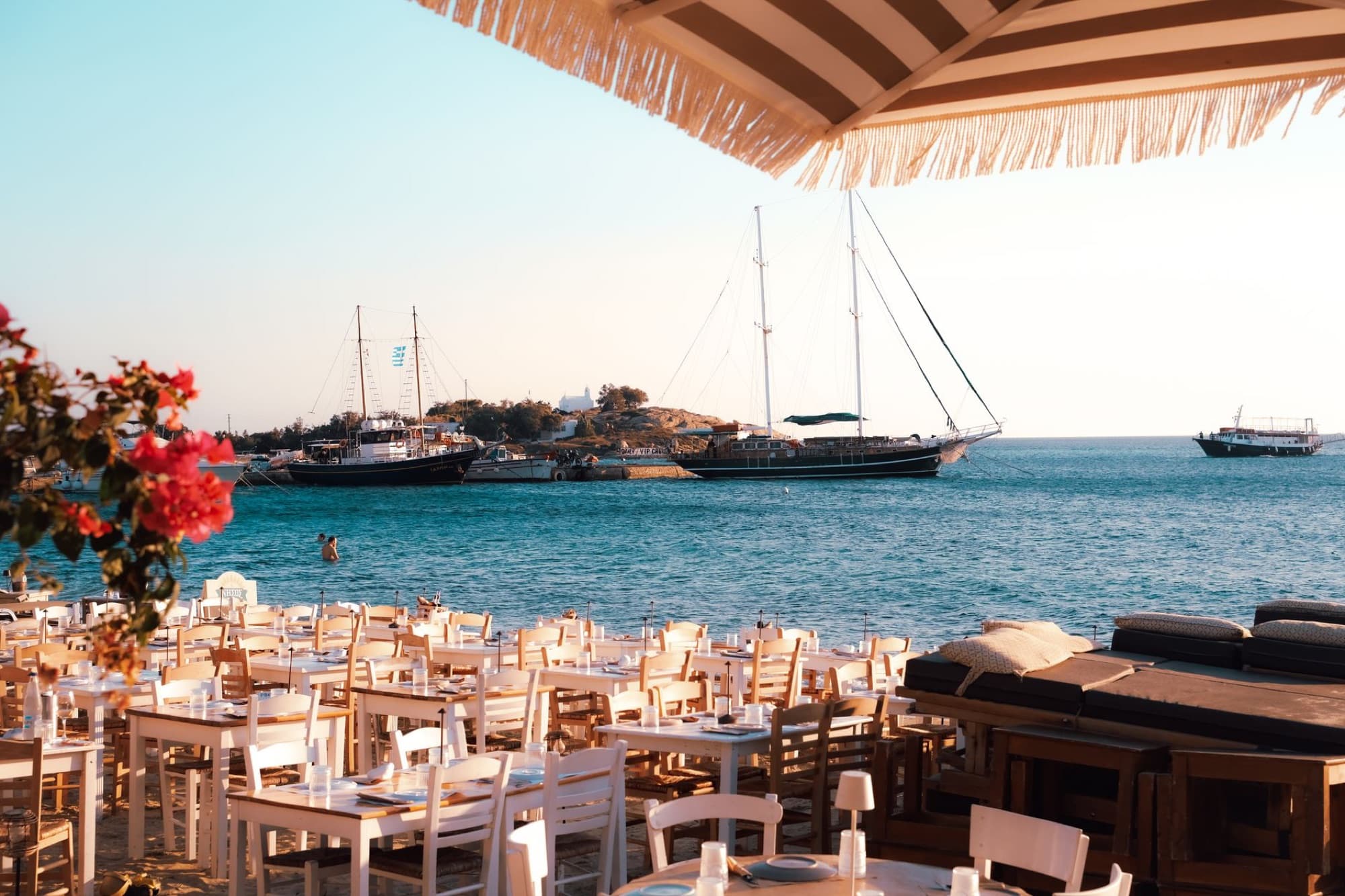 Beautiful beachside dining scene in Naxos, Greece with sailboats and clear blue waters at sunset.
