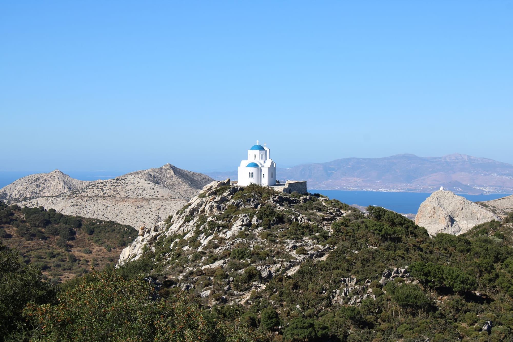 Picturesque white church with blue dome on a hill in Naxos, Greece, with sea and mountain views.