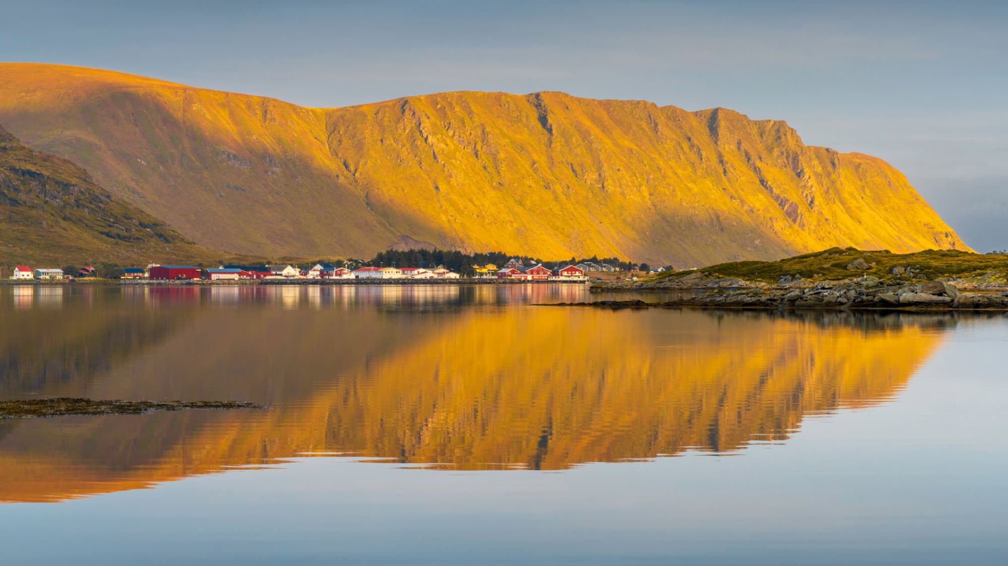 Scenic view of a coastal village reflecting in calm waters under golden mountains in Lofoten, Norway.