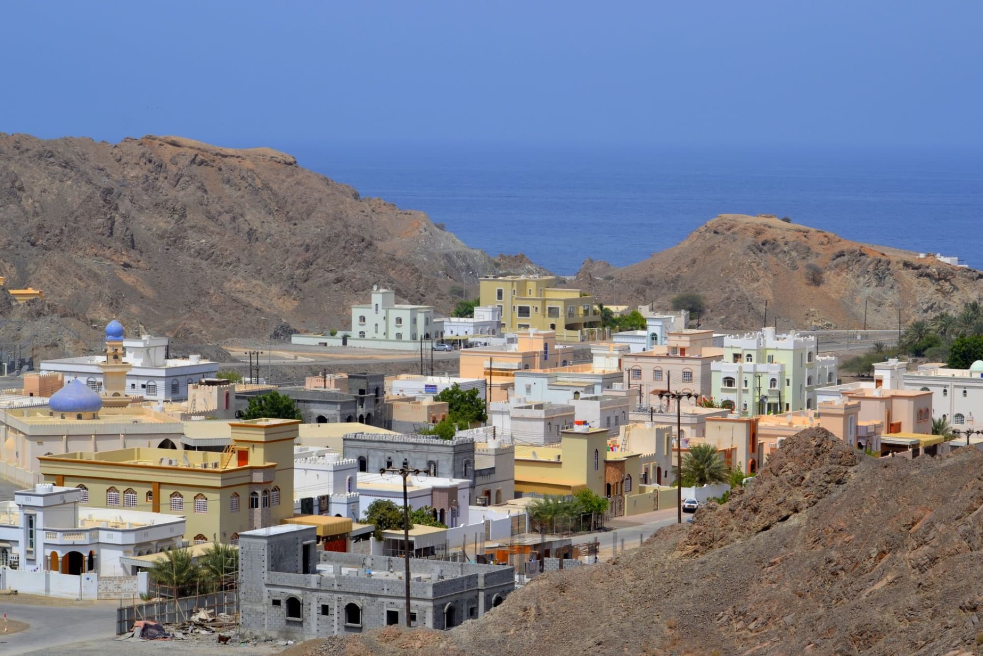 Scenic view of colorful architecture in a coastal town in Muscat, Oman, against arid hills and the sea.