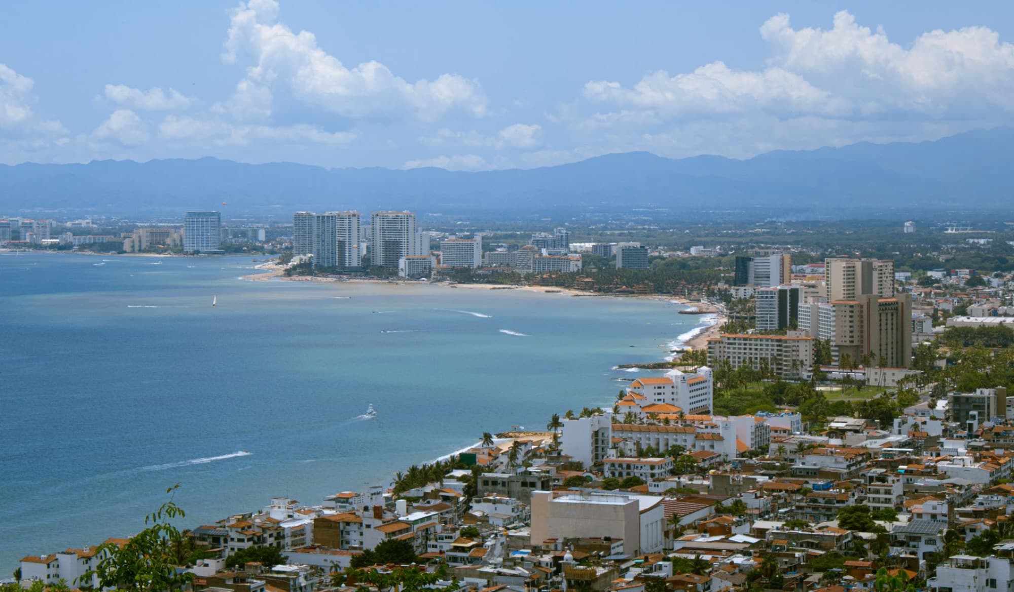 A panoramic cityscape of Puerto Vallarta, showcasing the beach, skyline, and marina against a mountain backdrop.