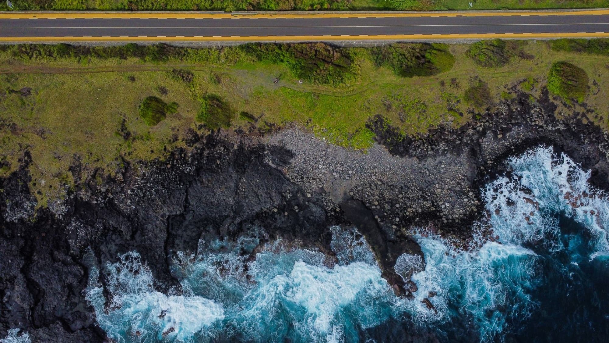 Drone view of La Réunion's coast featuring a road, rocky shoreline, and waves crashing on the beach.