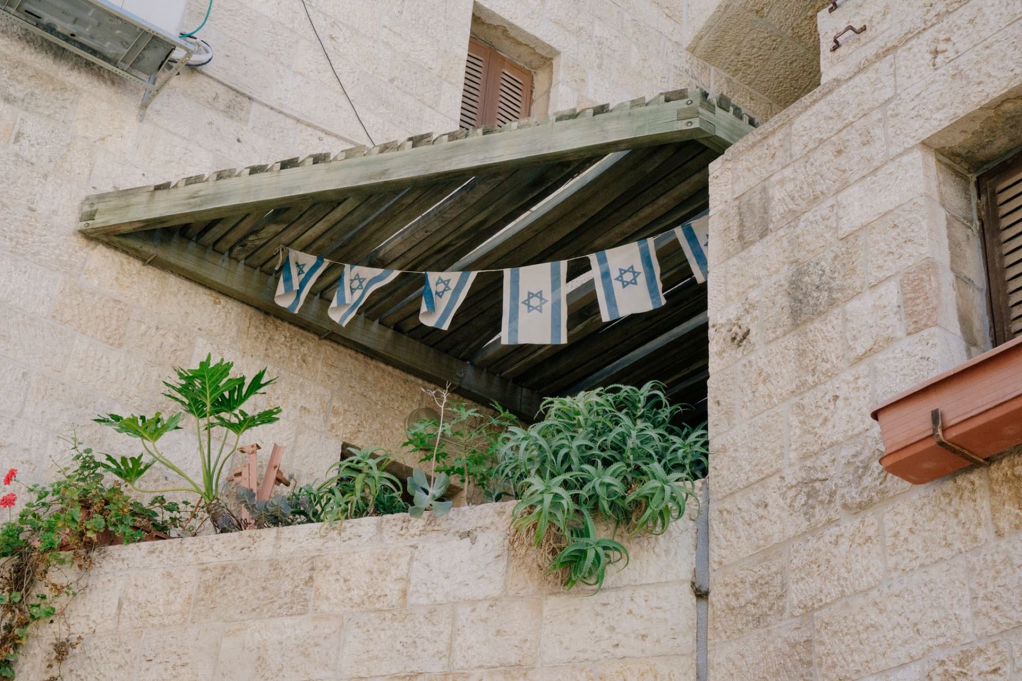 Stone building exterior with Israeli flags and diverse plants on balcony in urban setting.