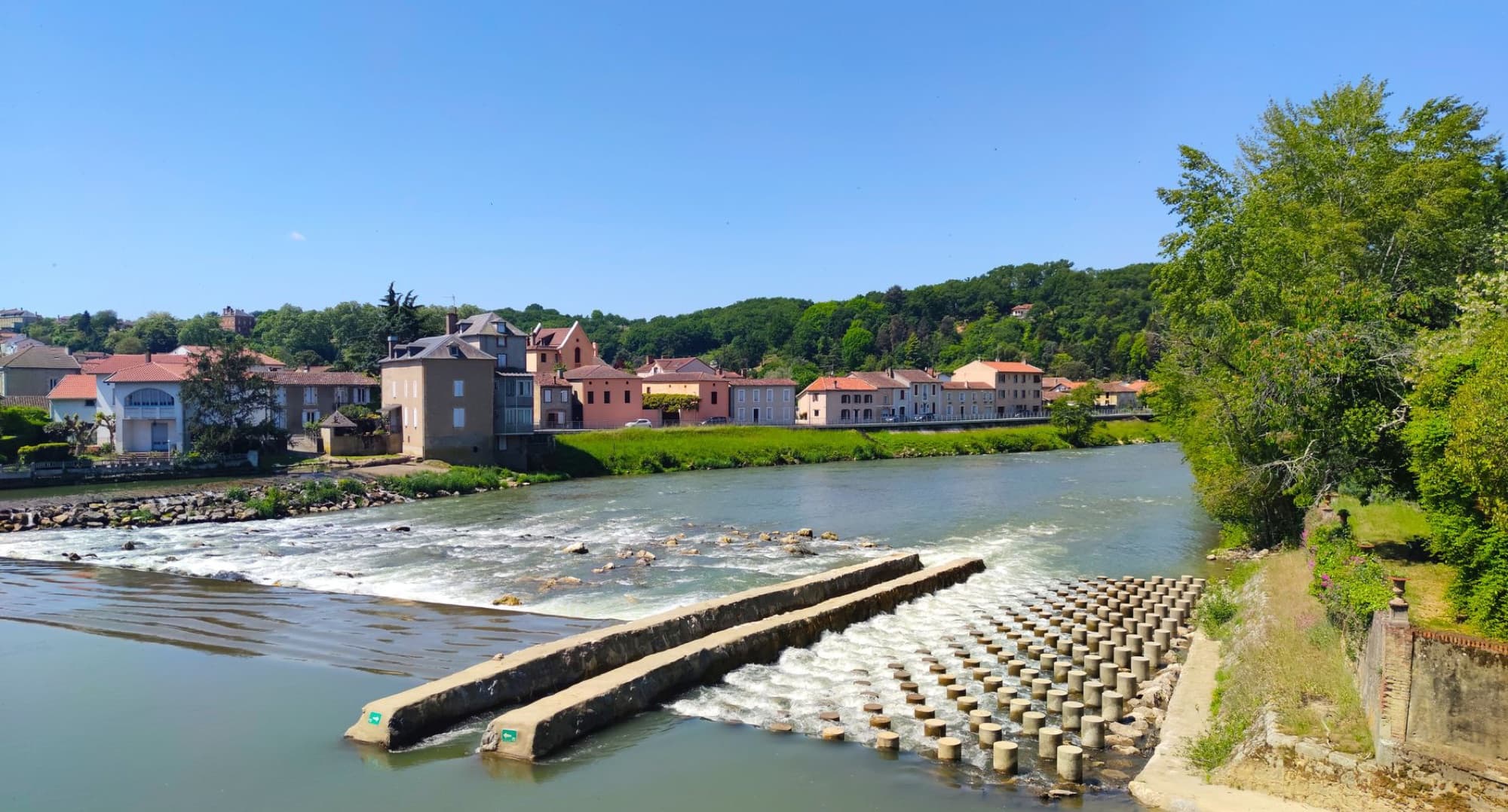 A scenic view of Aire-sur-l'Adour with colorful houses along the river under a clear blue sky.