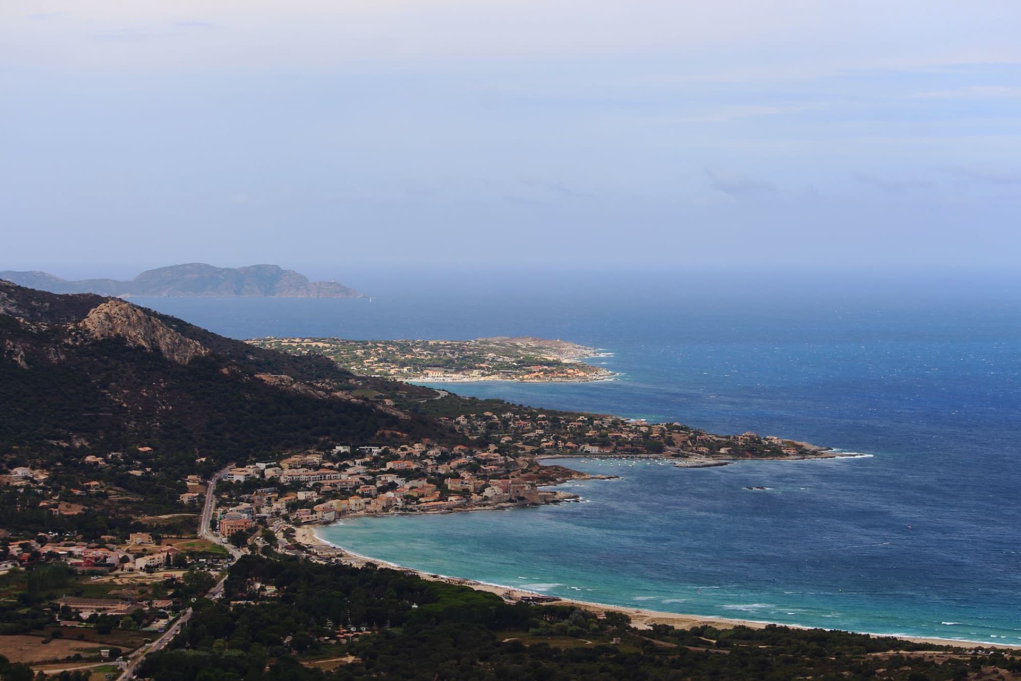 Drone view of coastal city located near hill on grassy coast near rippling sea against blue sky in tropical resort