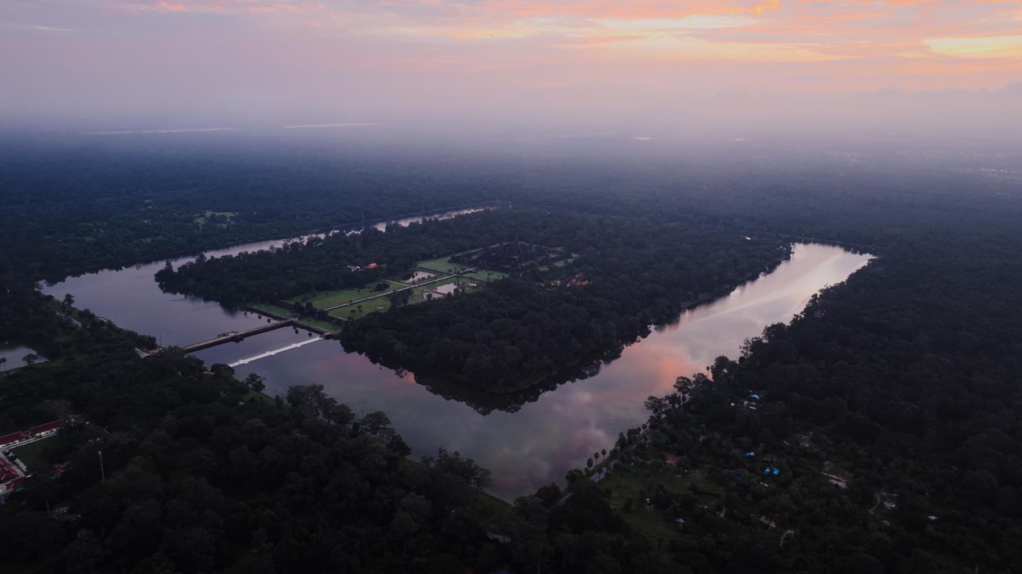 A breathtaking aerial view of Angkor Wat surrounded by lush forest at sunset, reflecting water bodies in Krong Siem Reap, Cambodia.