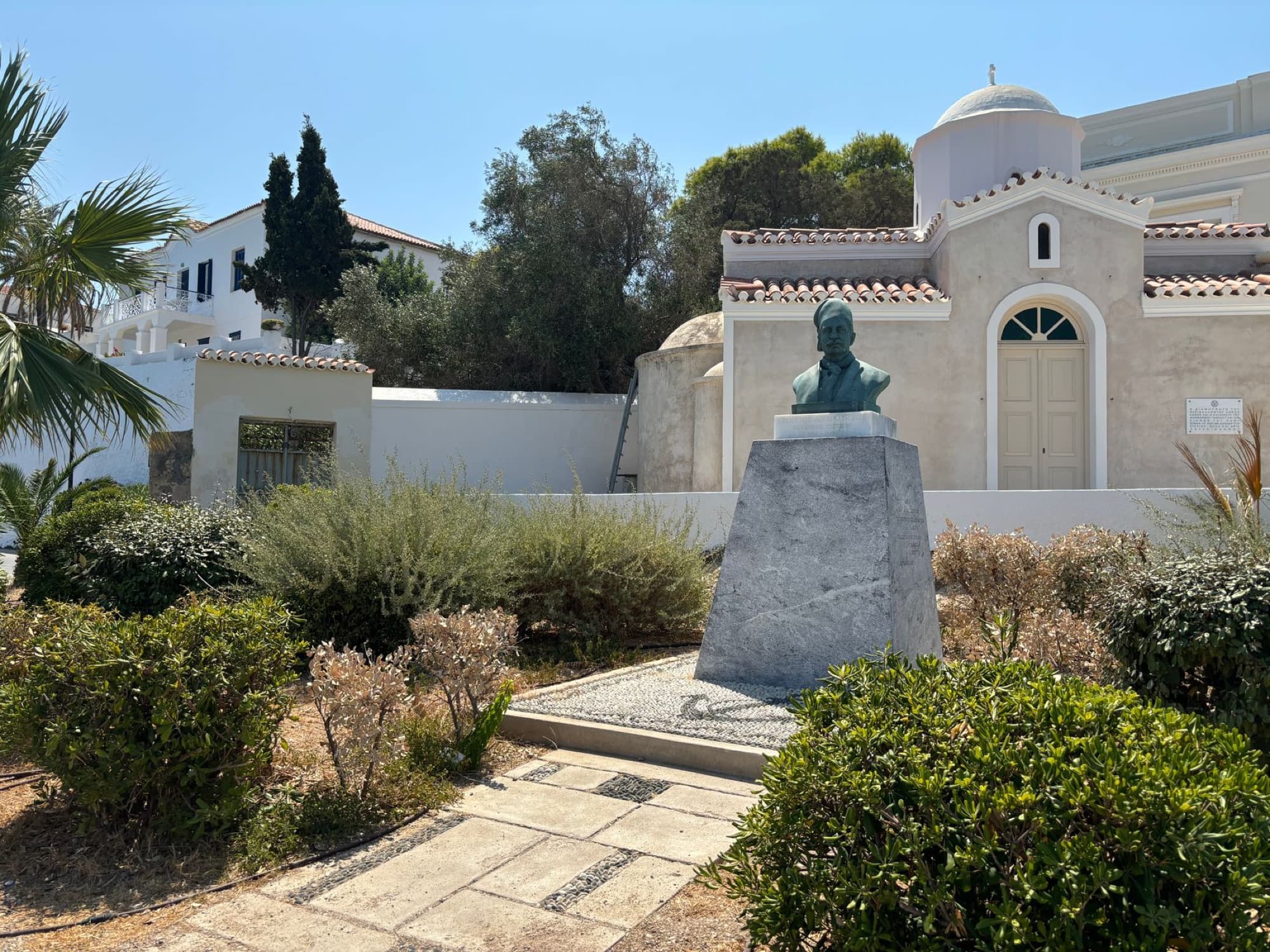 A bronze bust in front of an Orthodox church on a sunny day in Spetses, Greece.