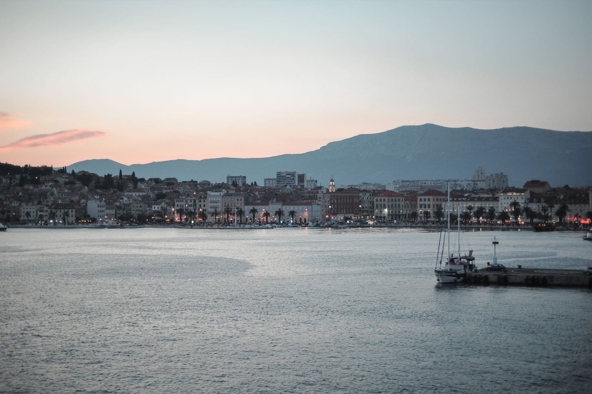 Picturesque view of river water with sailboat near modern town and huge mountain at sunset