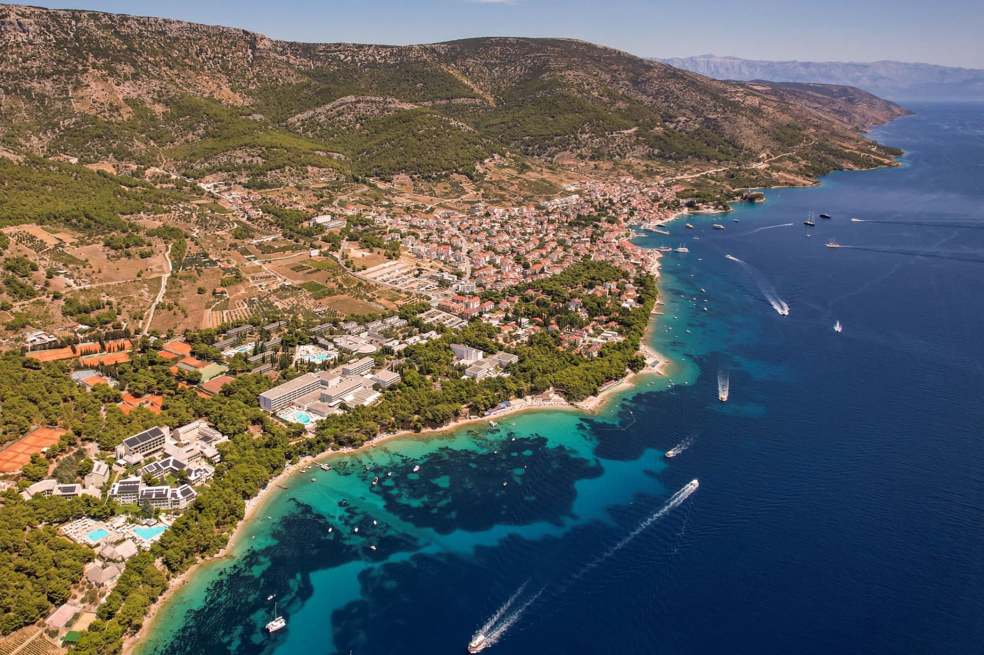 Aerial view of a coastal town with boats near a scenic shoreline and lush hillsides under a clear sky.