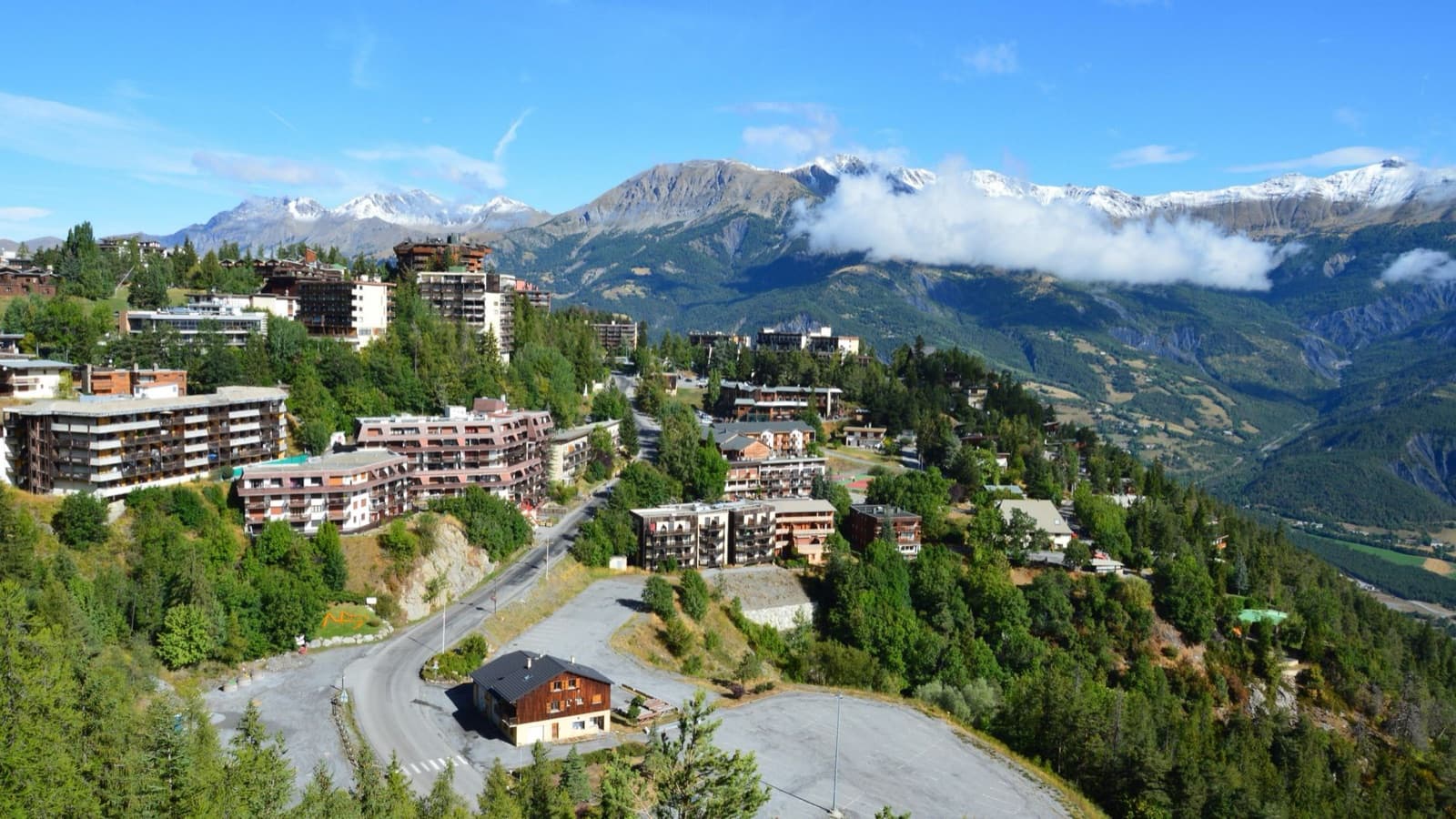 Scenic aerial view of a mountain village with buildings nestled within lush greenery and stunning alpine background.