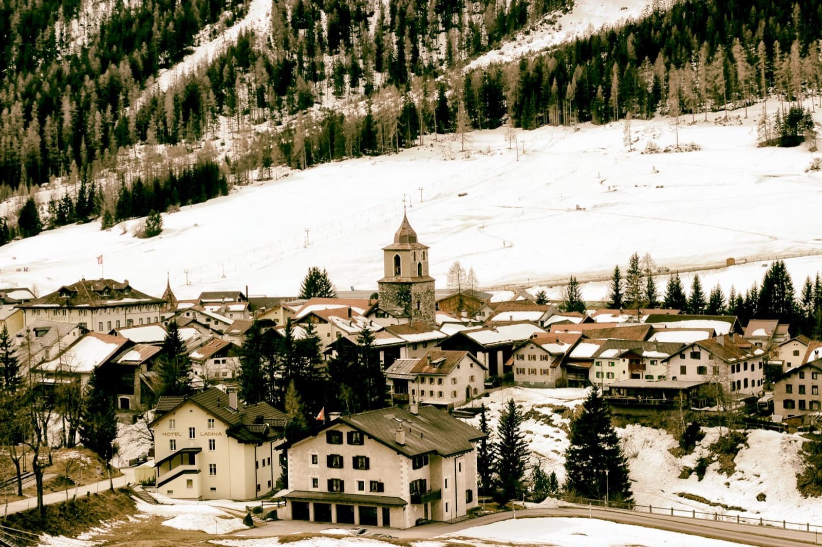 Scenic aerial view of a snowy Alpine village surrounded by mountains and trees in winter.