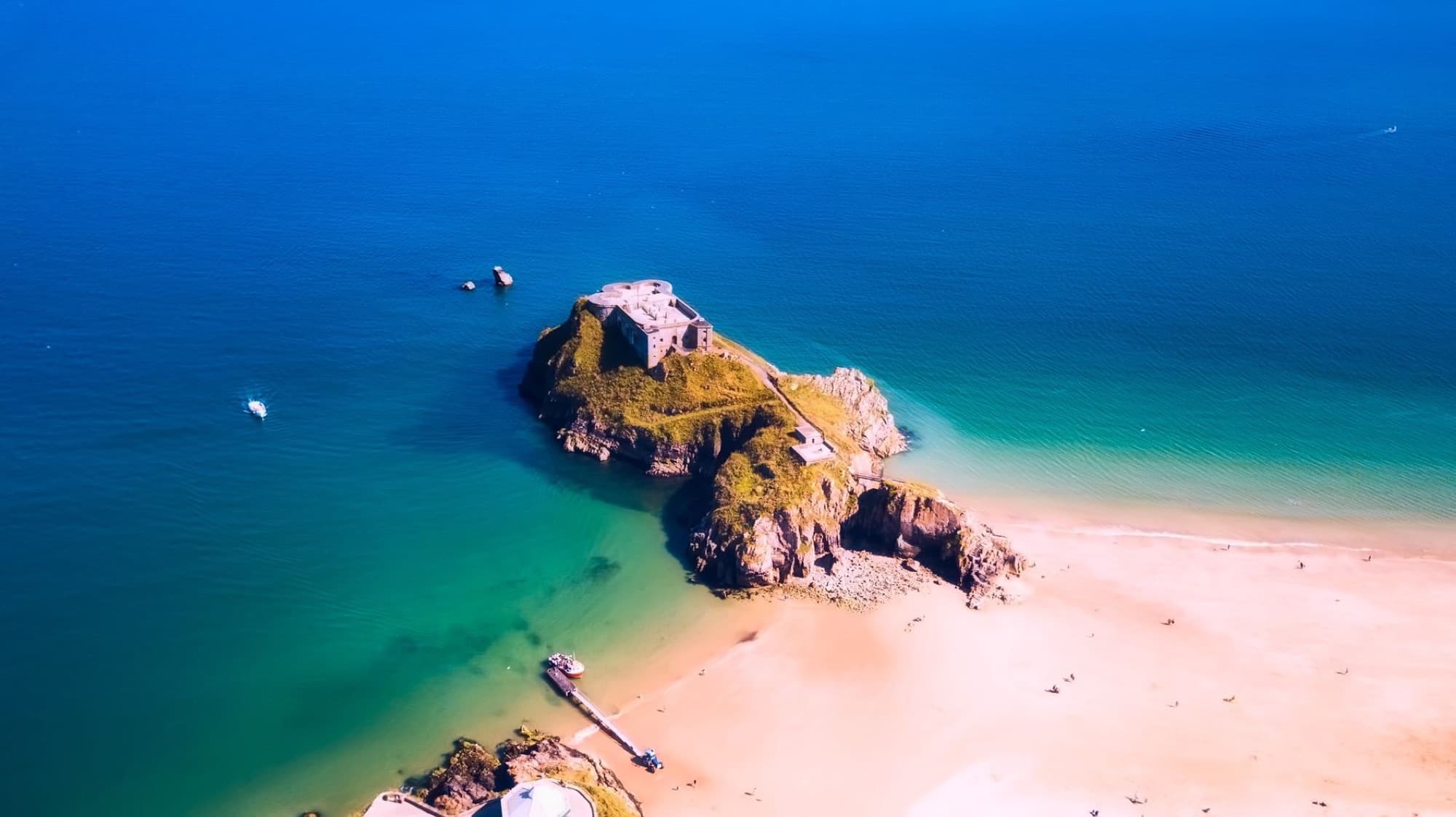 Aerial shot of a rocky island with ruins, surrounded by vibrant blue sea and sandy beach.