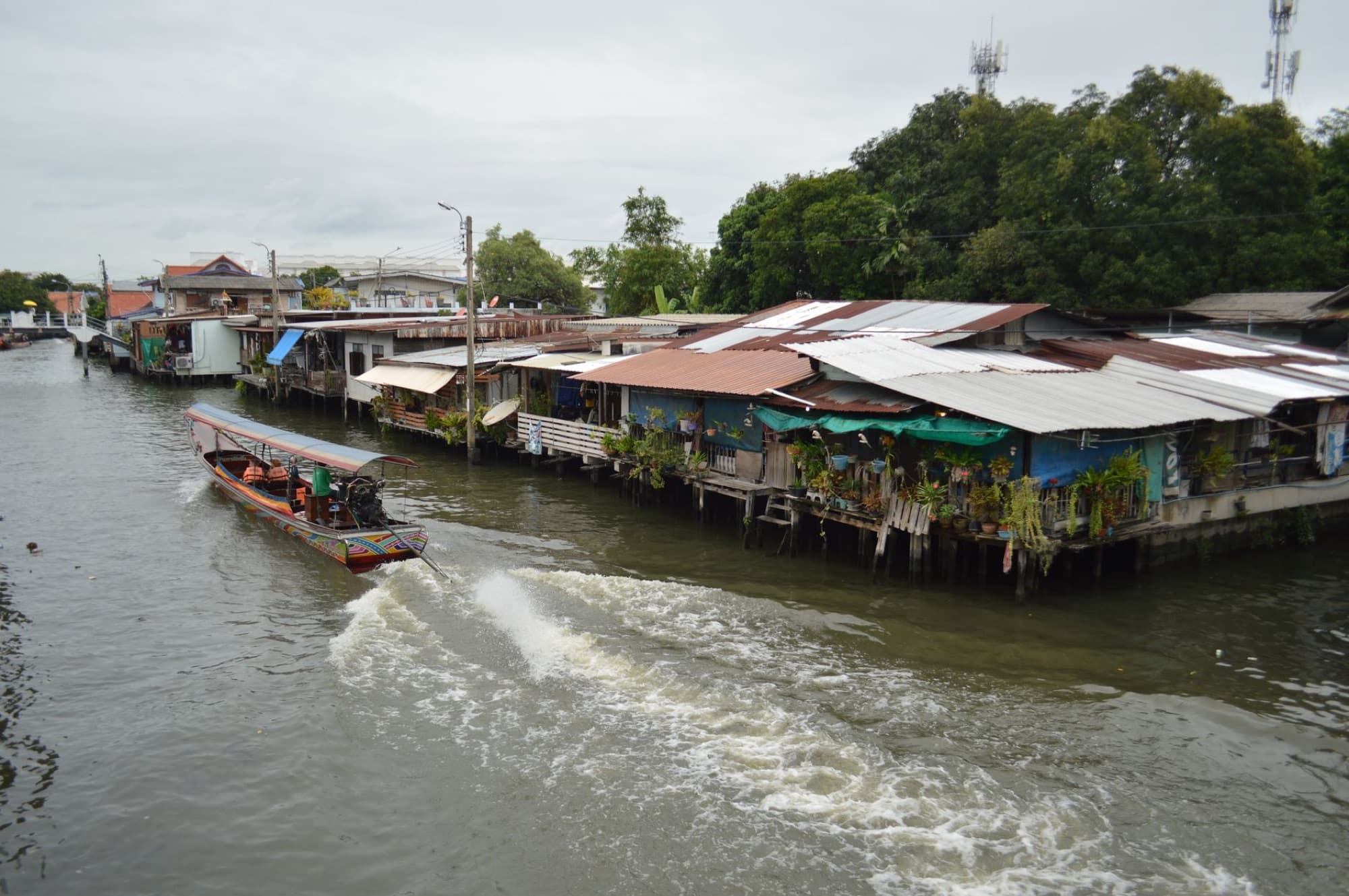 A scenic view of a traditional wooden boat navigating a canal beside rustic waterfront houses in Thailand.