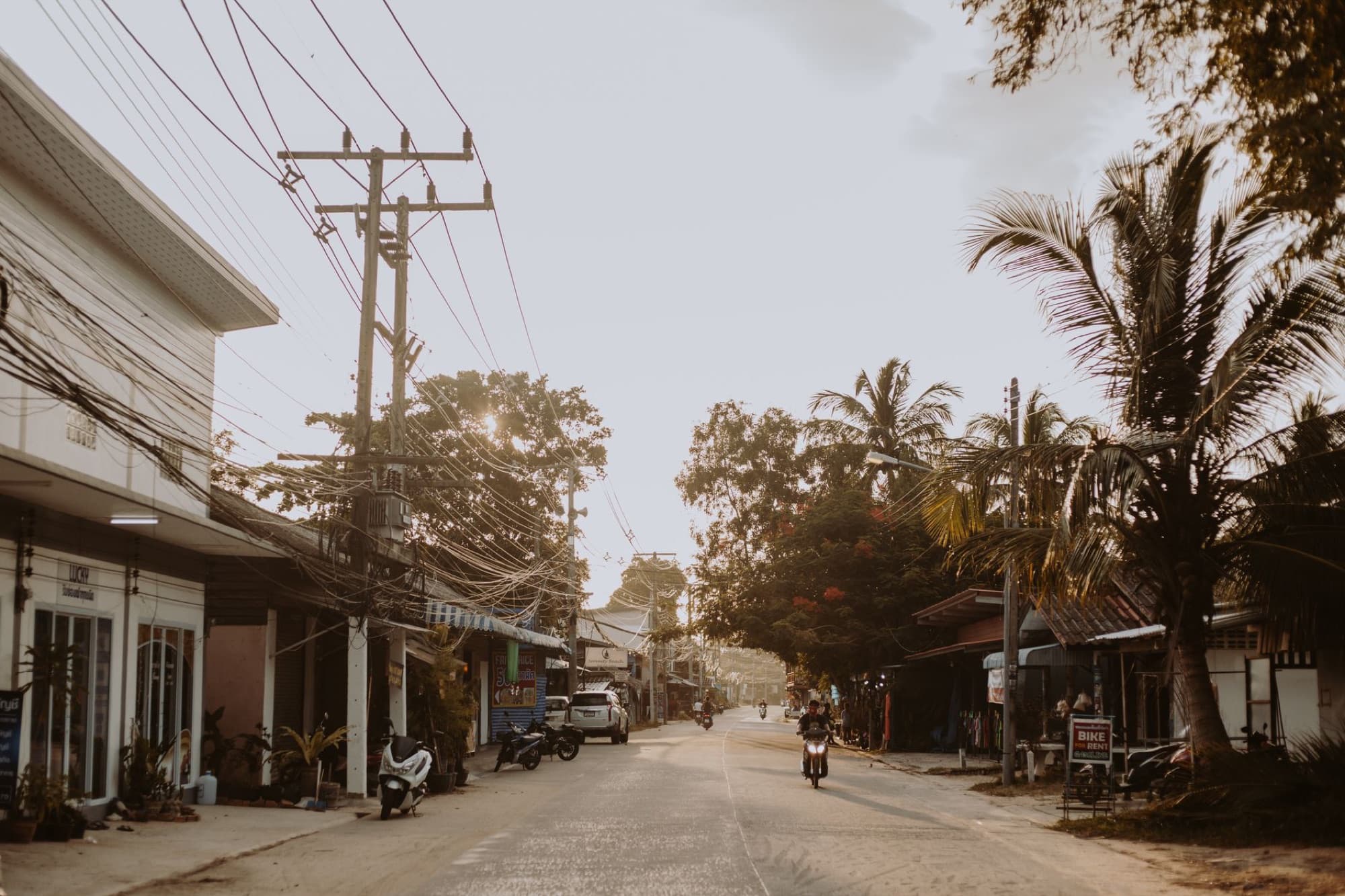 A peaceful urban street in Thailand with motorcycles and tropical trees under a bright sky.