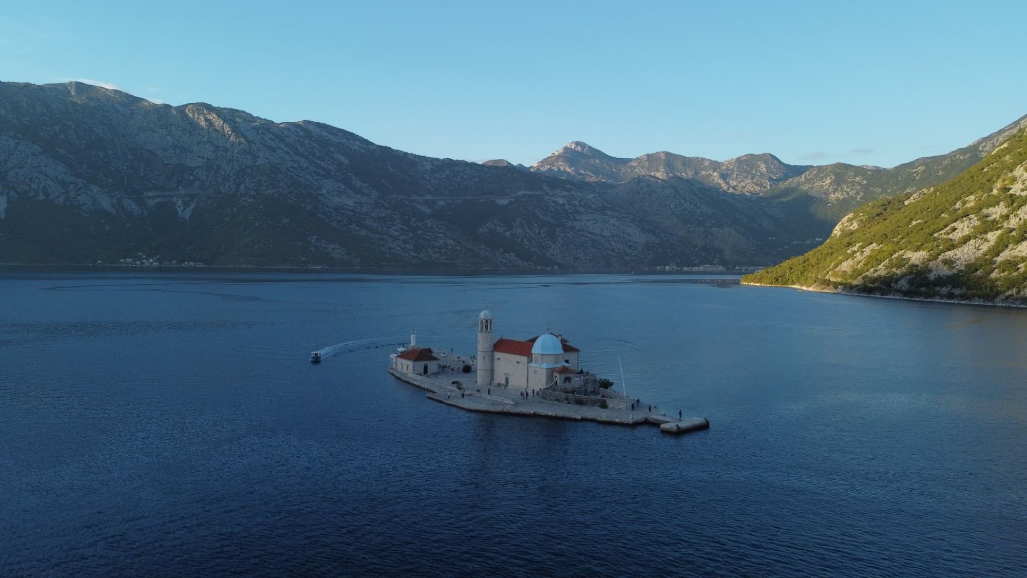 Aerial view of Our Lady of the Rocks island in Kotor Bay, Montenegro, surrounded by tranquil waters and mountains.