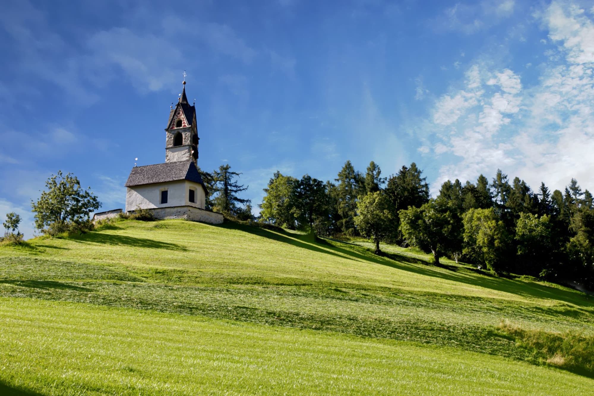 Picturesque church perched on a green hillside in Trentino-Alto Adige, surrounded by nature under a blue sky.