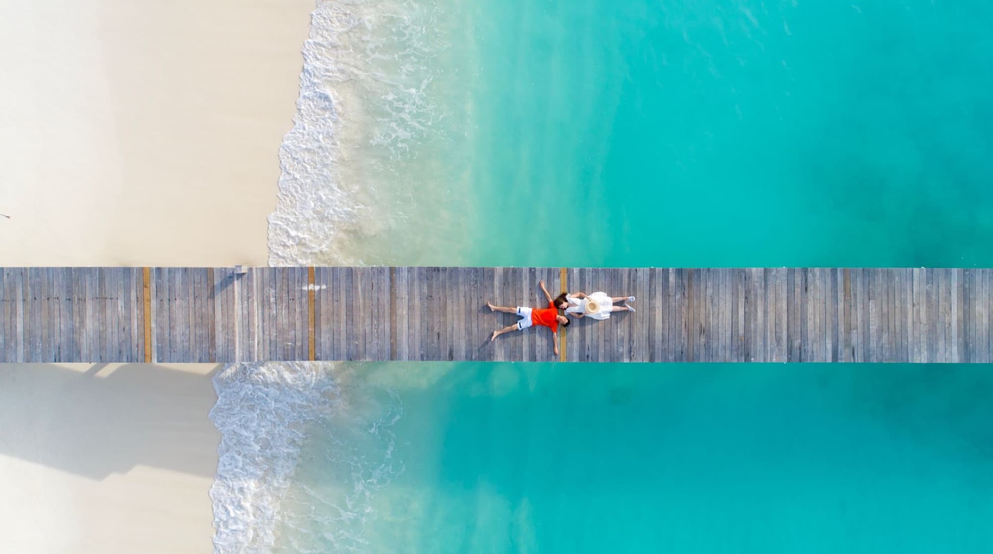 A serene aerial shot of a couple lying on a wooden dock over clear blue ocean by a sandy beach.