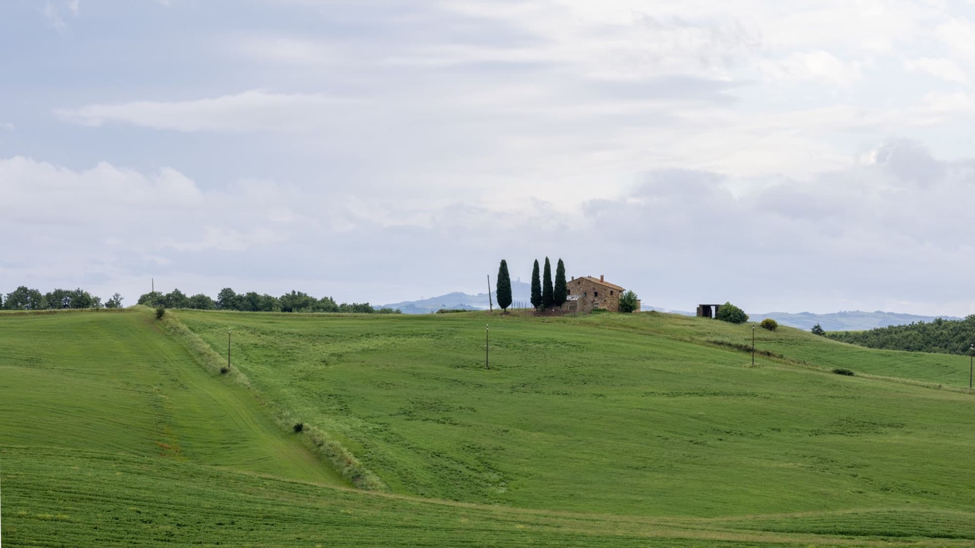 Scenic view of rolling green hills and a farmhouse in Pienza, Tuscany under a cloudy sky.