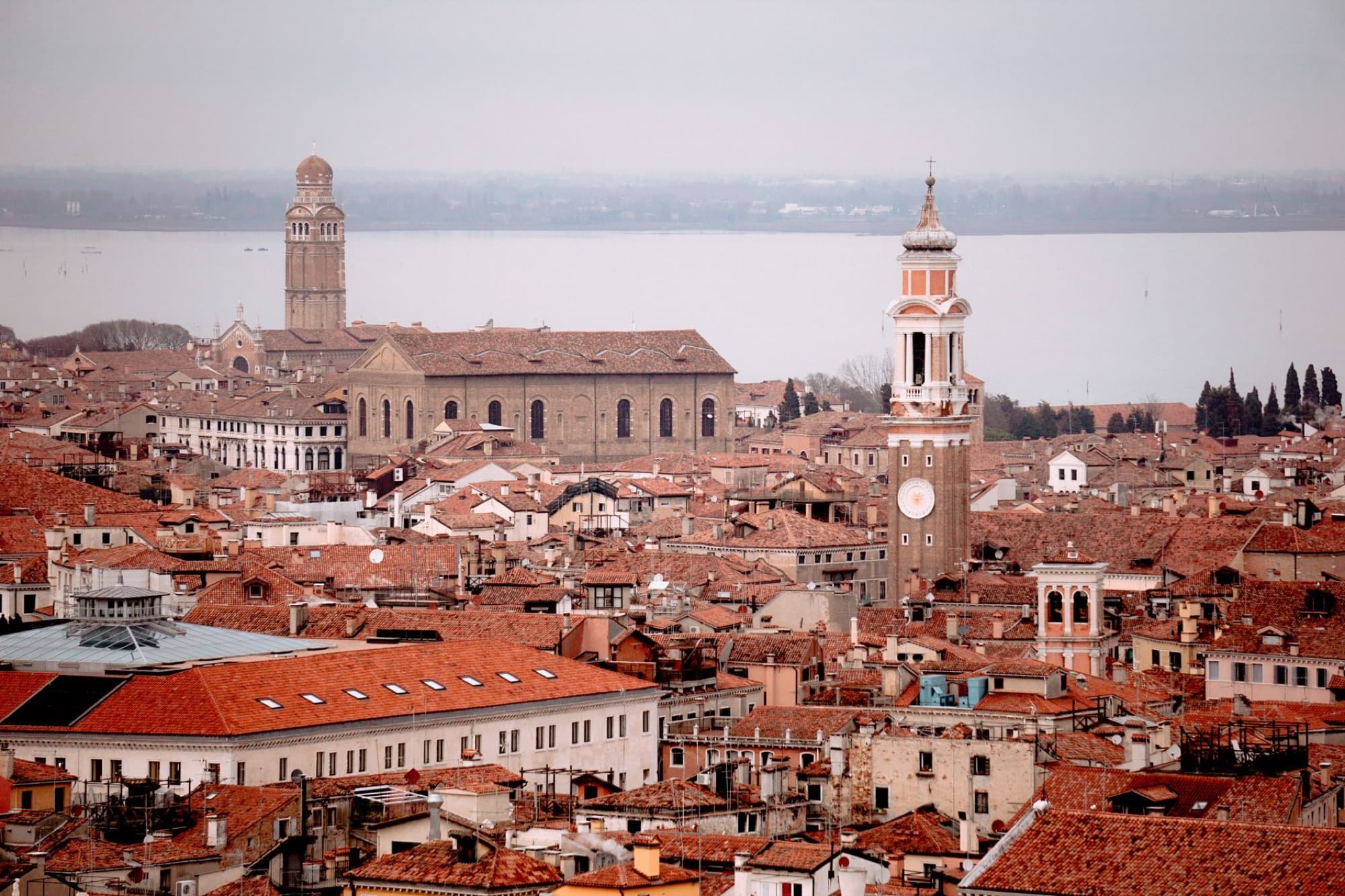 Panoramic aerial view of Venice showcasing red rooftops, towers, and historic architecture under a cloudy sky.