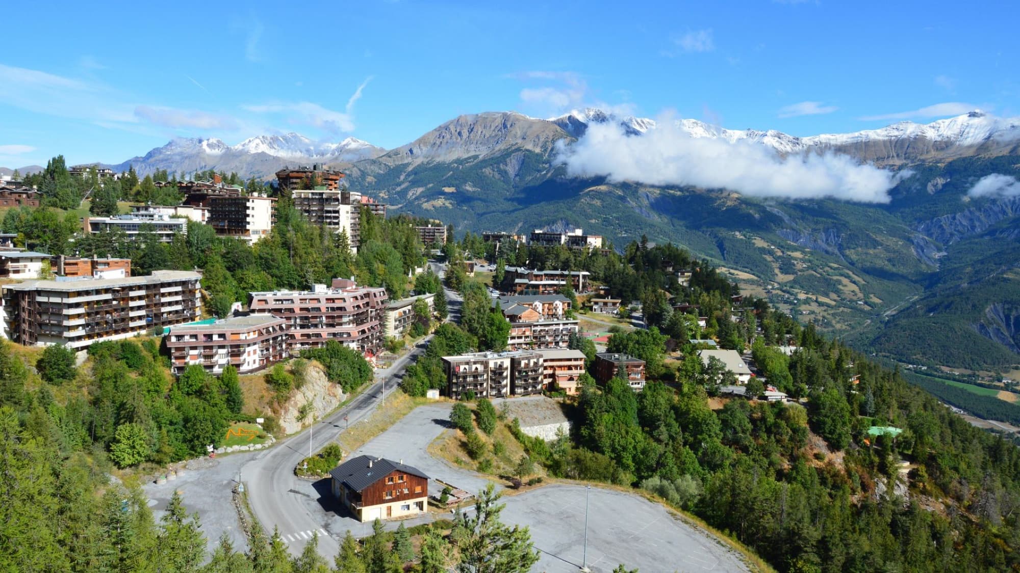 Scenic aerial view of a mountain village with buildings nestled within lush greenery and stunning alpine background.