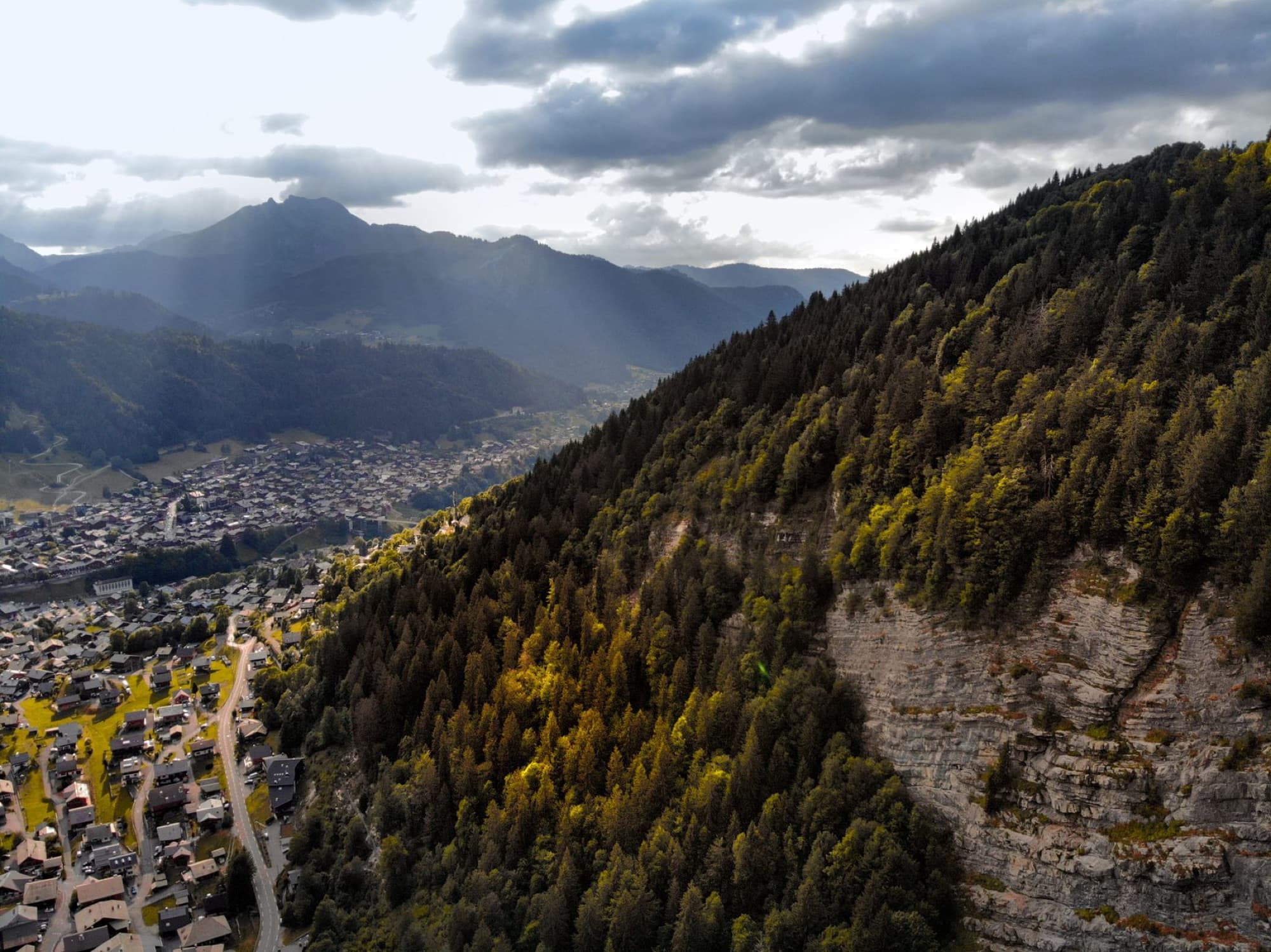 Drone captures a scenic view of a hillside town against the backdrop of mountains during evening light.
